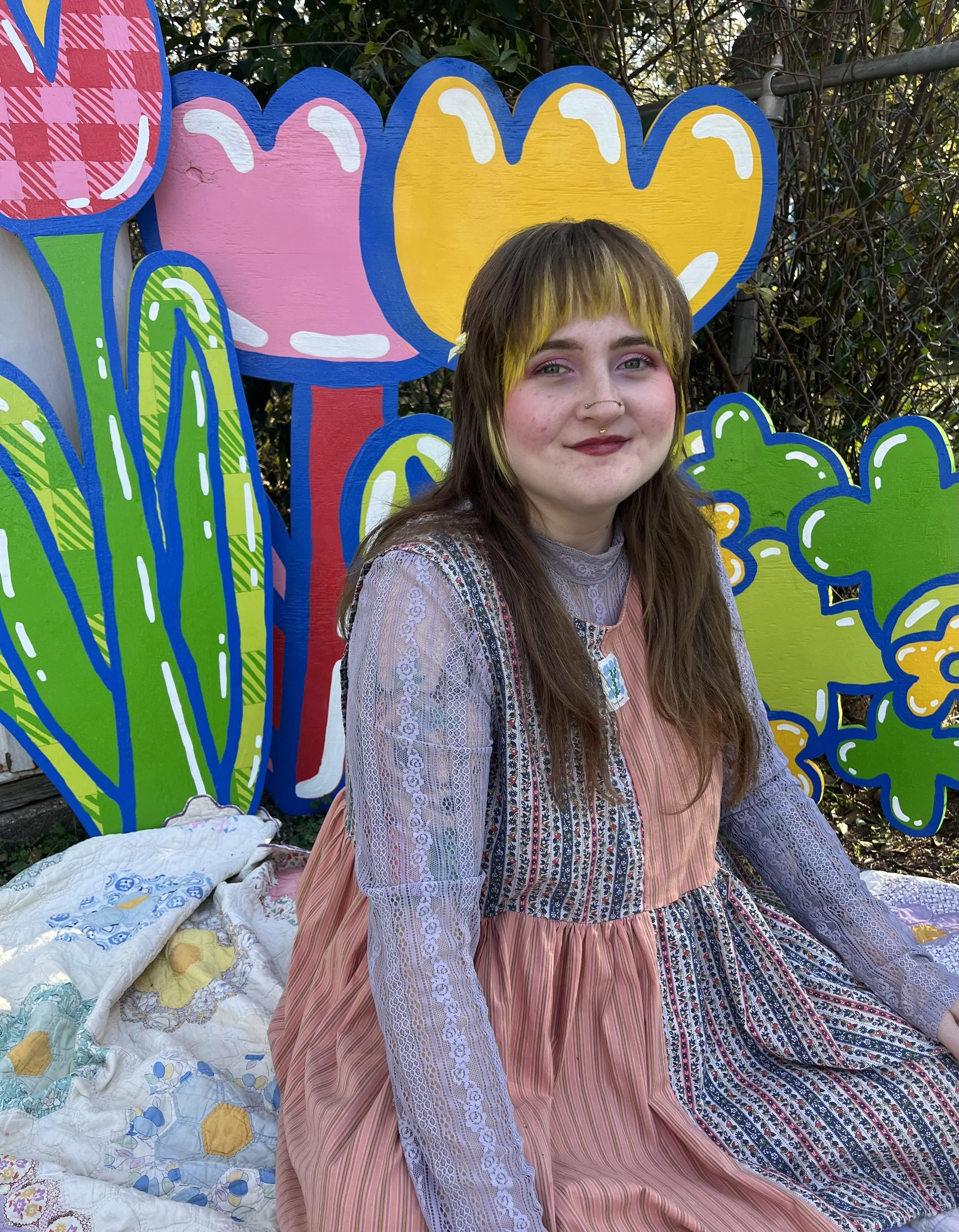A non-binary person wearing a color blocked dress sits in front of large cut-outs of hand painted flowers