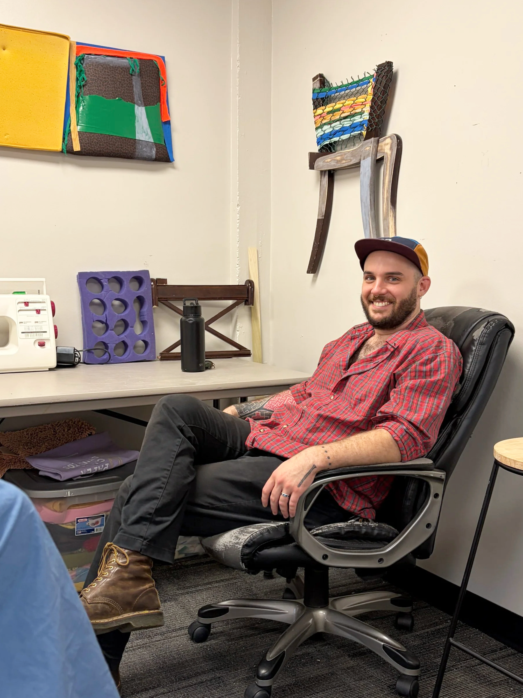 Artist Joshua Bryant sits in a chair at his work table in Back Door Studio with colorful artwork on the walls