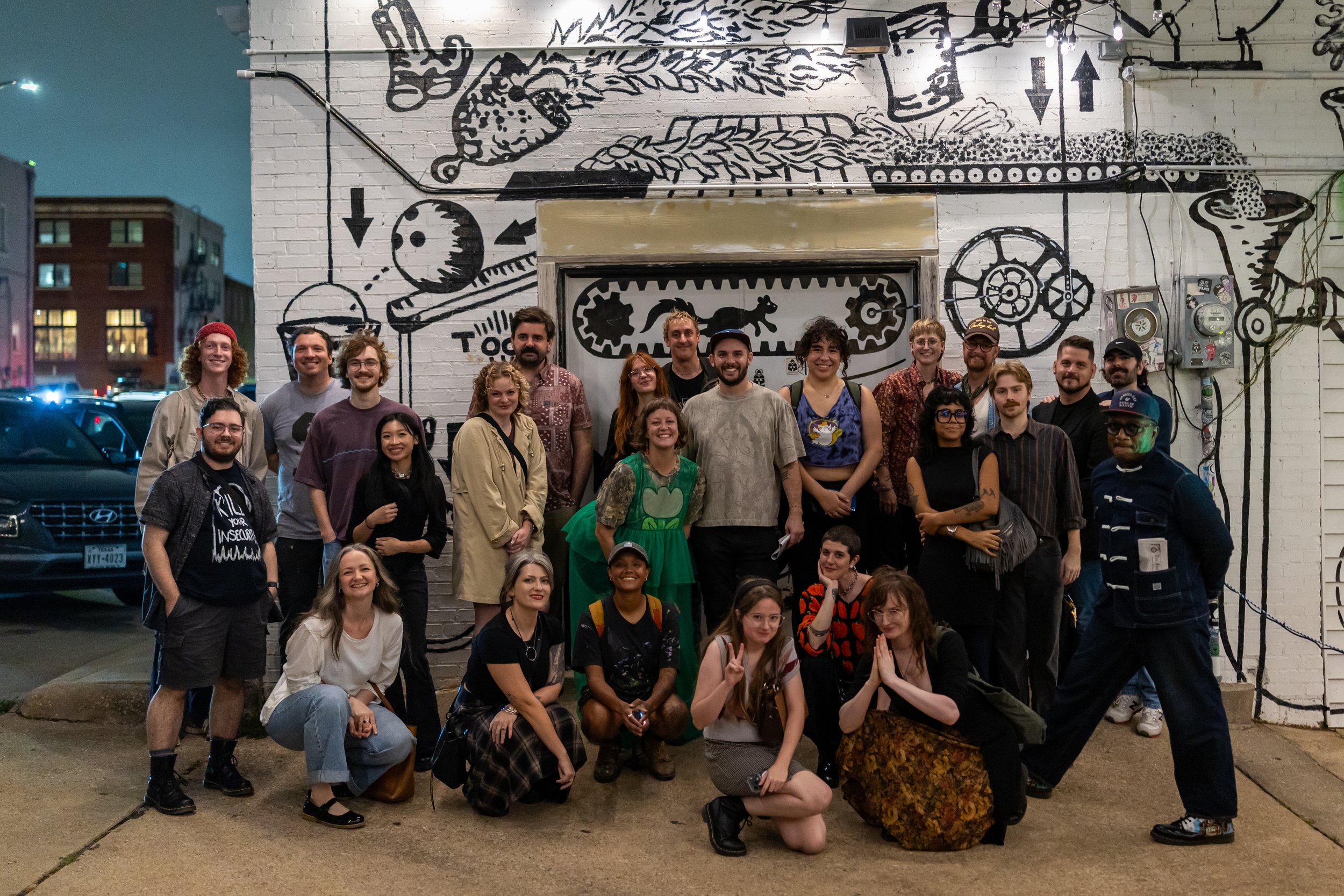 a group of 23 artists pose together in front of the black and white graphic mural at Bearded Monk