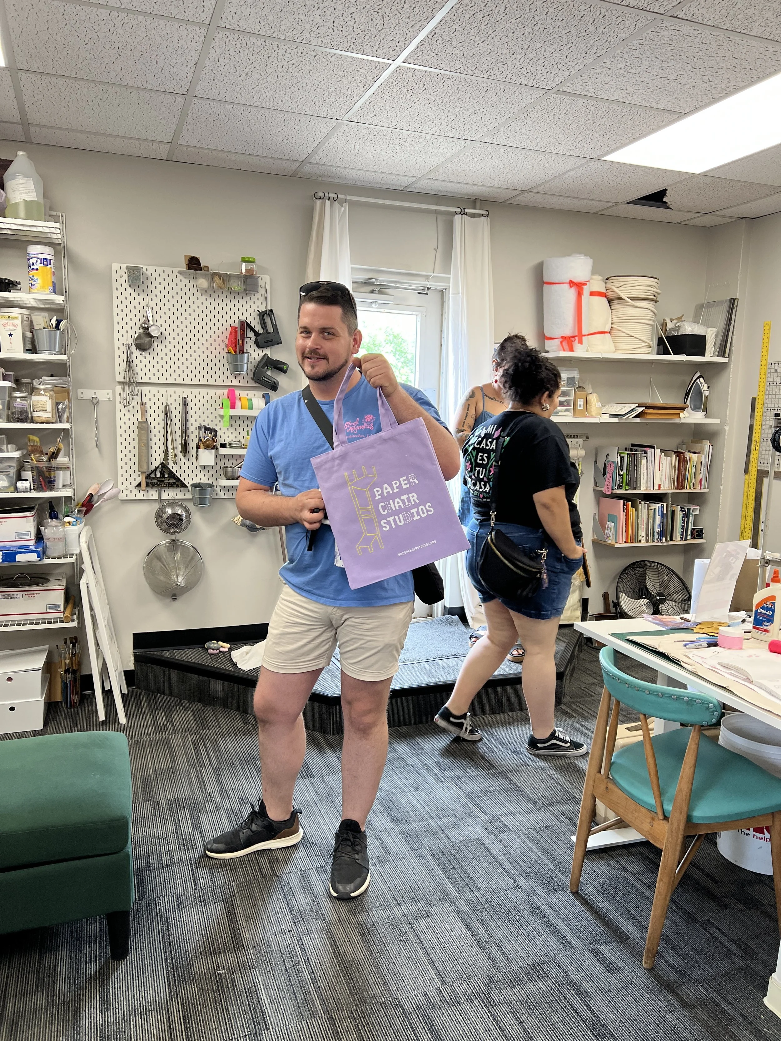 A tall white man holds a lavender tote with the Paper Chair Studios logo on it and is standing in a studio with shelves of paper equipment and other tools on the wall