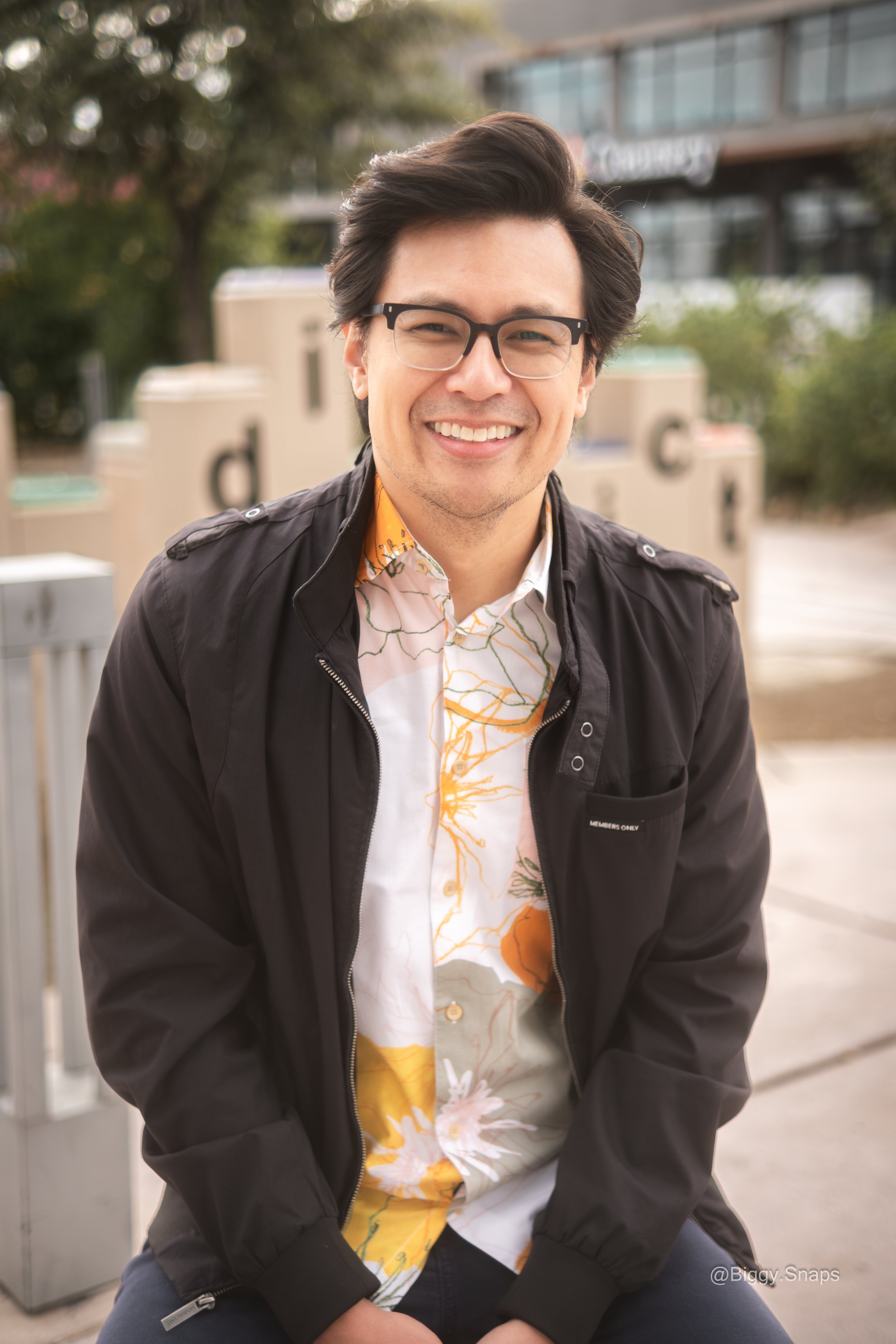 A young man with glasses and dark hair smiling outdoors, wearing a black jacket over a floral shirt, with trees and some white crates in the background.