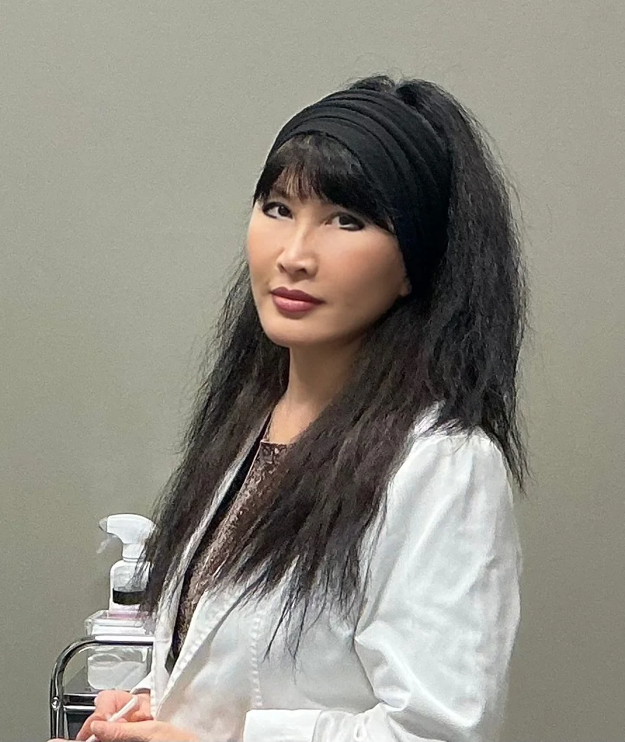 A woman with long, wavy black hair wearing a black headband and a white lab coat, standing in front of a neutral background.