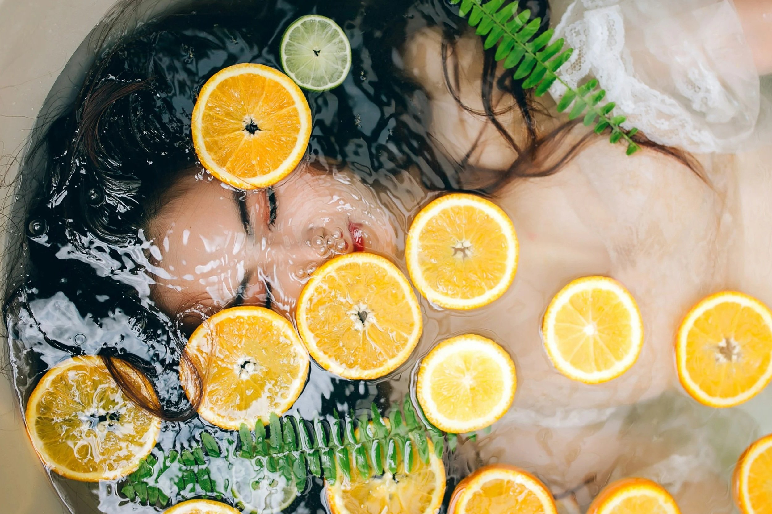 Woman relaxing in a bubble bath with orange and lime slices floating on water, a green fern leaf, and a white lace towel.