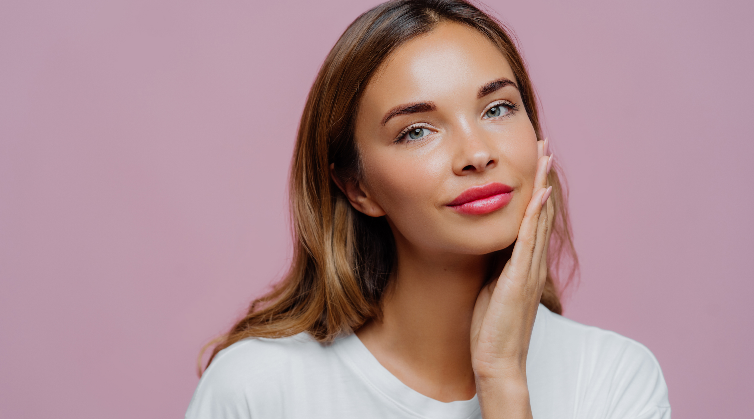 A woman with light skin, long brown hair, and blue eyes, touching her face gently with one hand, standing against a pink background.