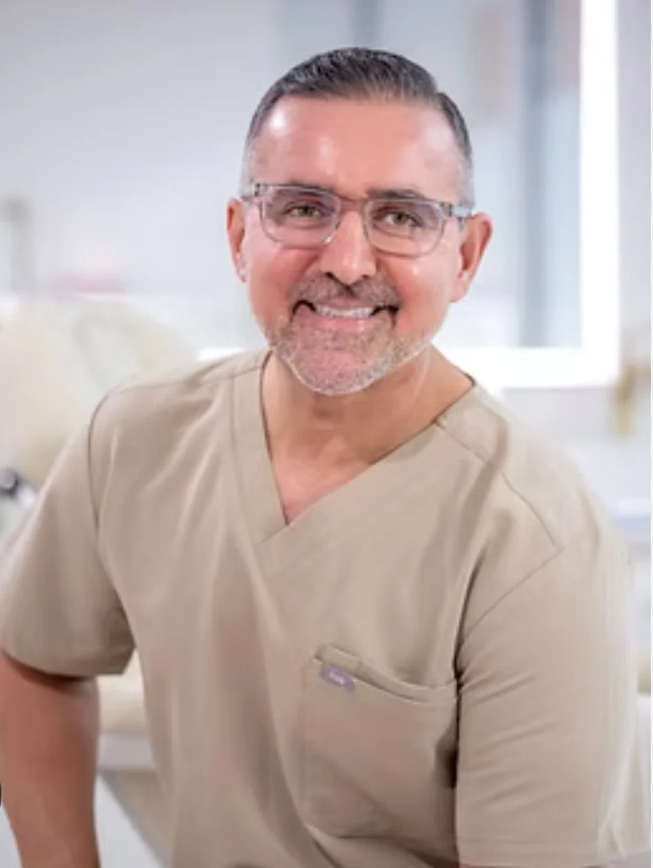 A smiling man with glasses, gray hair, and a beard, wearing beige medical scrubs, in a clinical setting.
