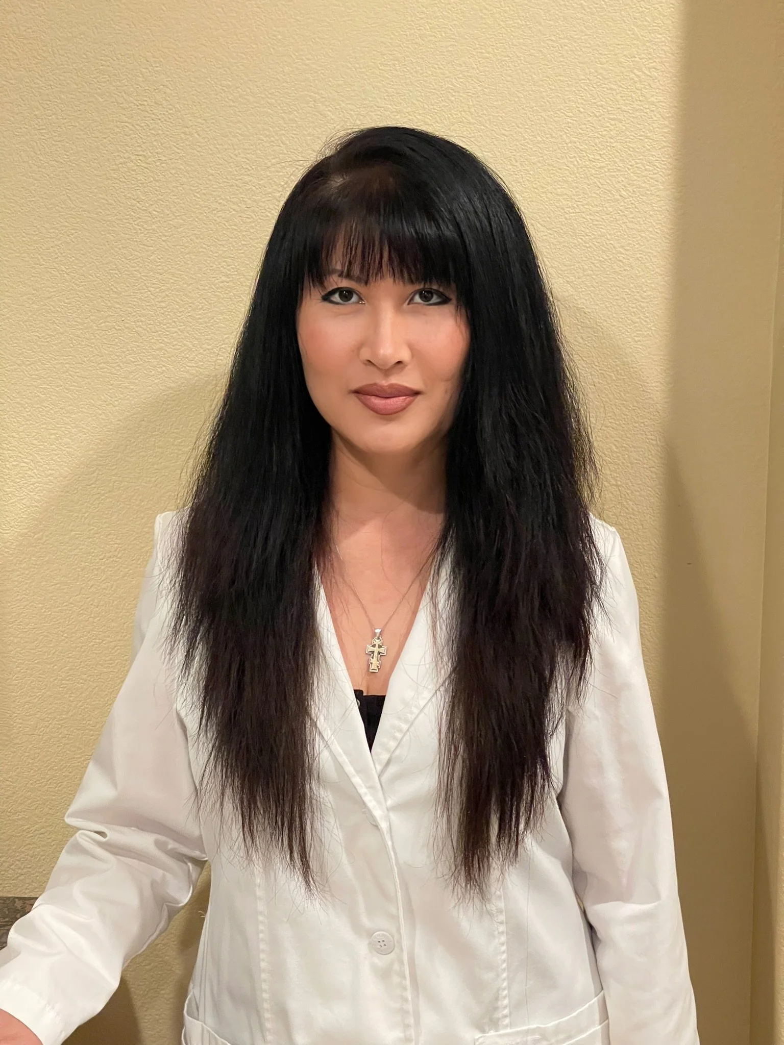 A woman with long black hair and bangs wearing a white coat and a cross necklace standing against a beige wall.