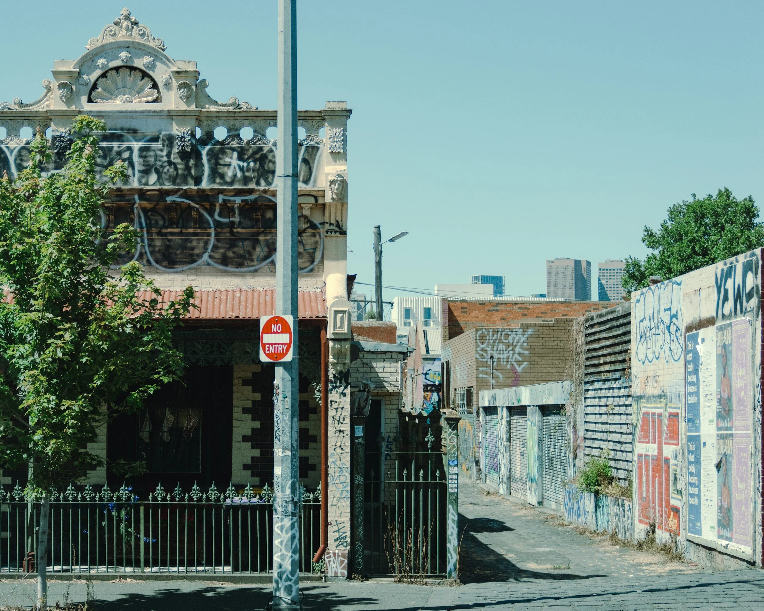 Street scene with graffiti-covered buildings, no entry sign, trees, and a partly visible old building with decorative architecture in the background.