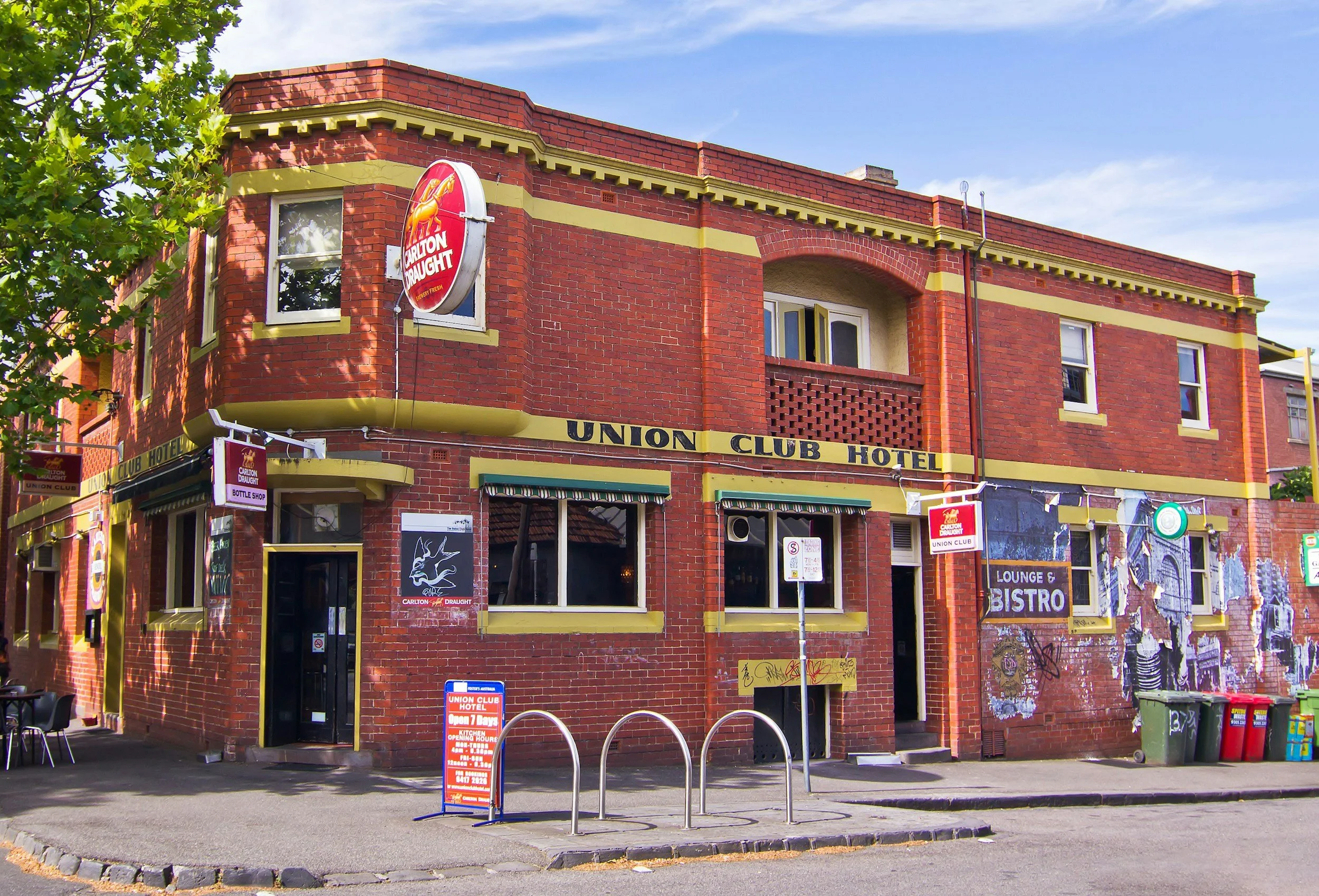 Red brick building labeled 'Union Club Hotel' with signs for lounge and bistro, Carlton Draught beer advertisement, and graffiti art on the walls. There are chairs and waste bins on the sidewalk.