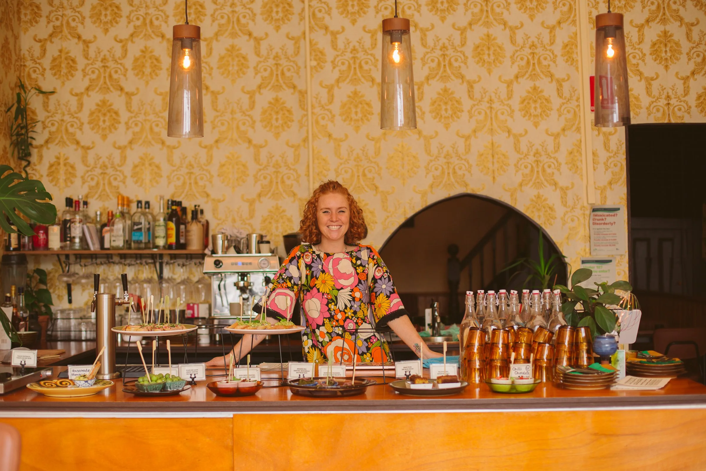 A cheerful woman with red curly hair and a colorful floral dress standing behind a bar counter with various drinks, glasses, and plates of food in an elegant, vintage-style restaurant with yellow damask wallpaper.