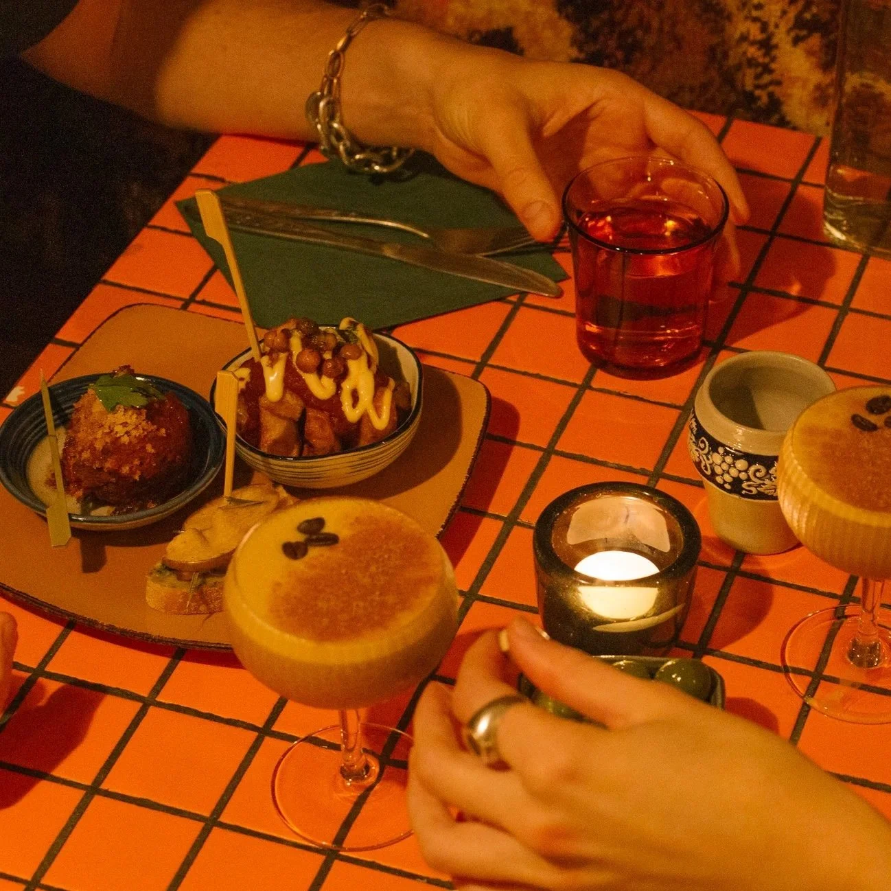 People enjoying drinks and small dishes on a tiled table in a dimly lit setting, with food, beverages, and a candle.