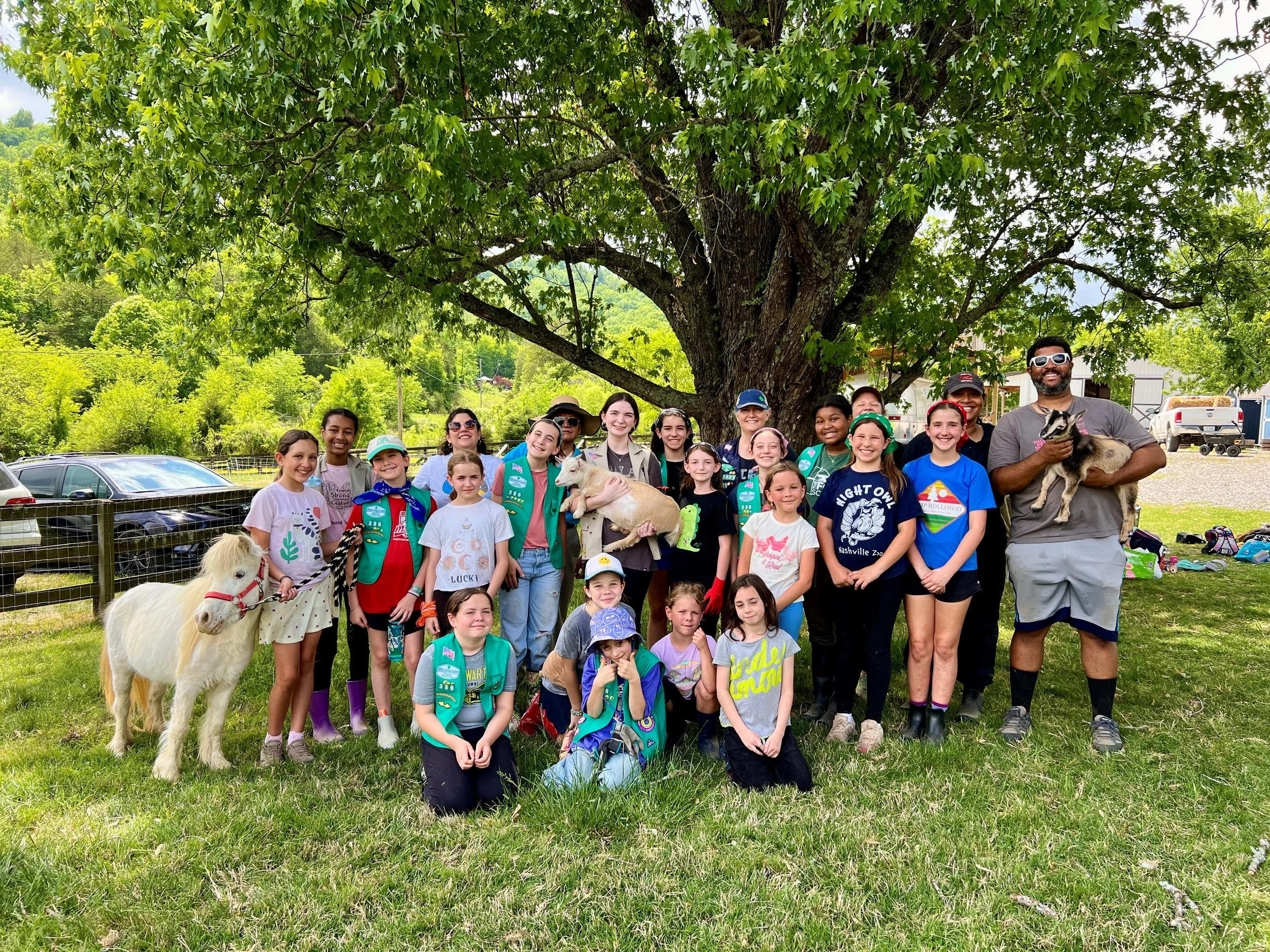 A large group of people, mostly children, gathered outdoors under a large leafy tree, some people holding goats and smiling, with a man holding a goat on the right.