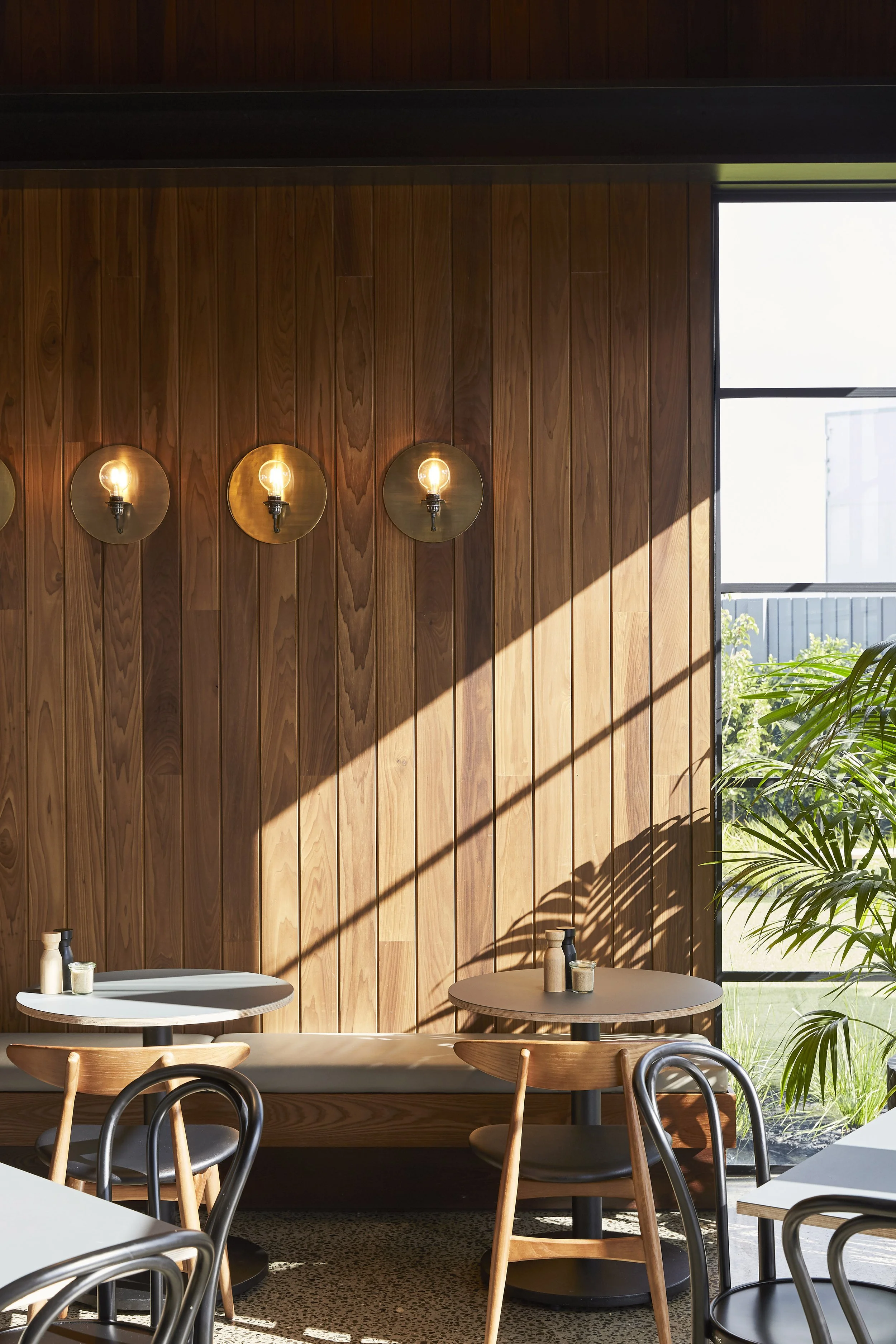 Interior of a modern cafe with wood-paneled walls, round wall sconces with bulbs, large windows letting in natural light, and small round tables with chairs. Green plants are visible outside.