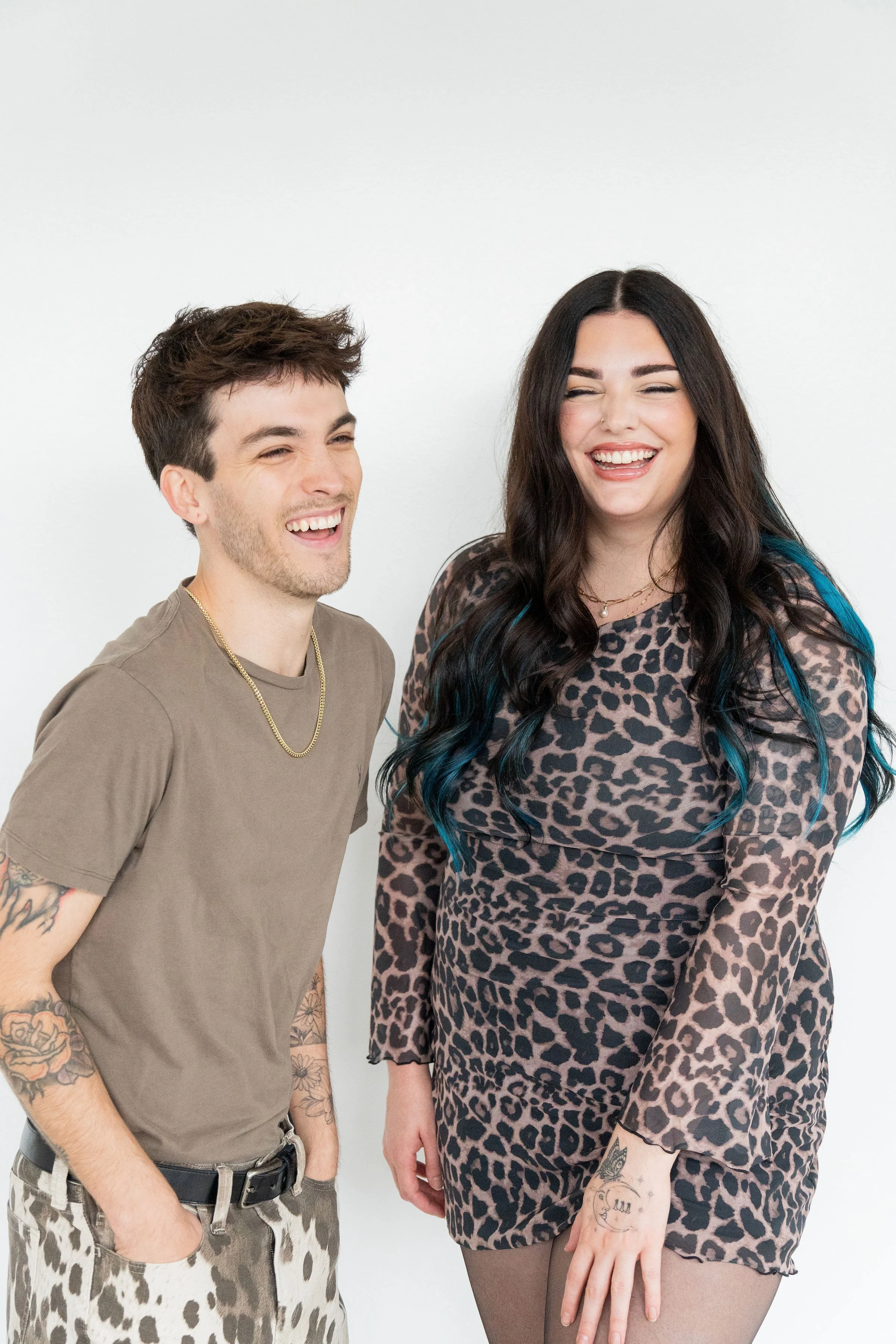 Two people, a man and a woman, are laughing and standing close together against a plain white background. The man has short brown hair, tattoos on his arms, and is wearing a beige t-shirt, leopard print pants, and a gold chain. The woman has long dark hair with blue streaks, multiple tattoos, and is wearing a leopard print dress and tights.