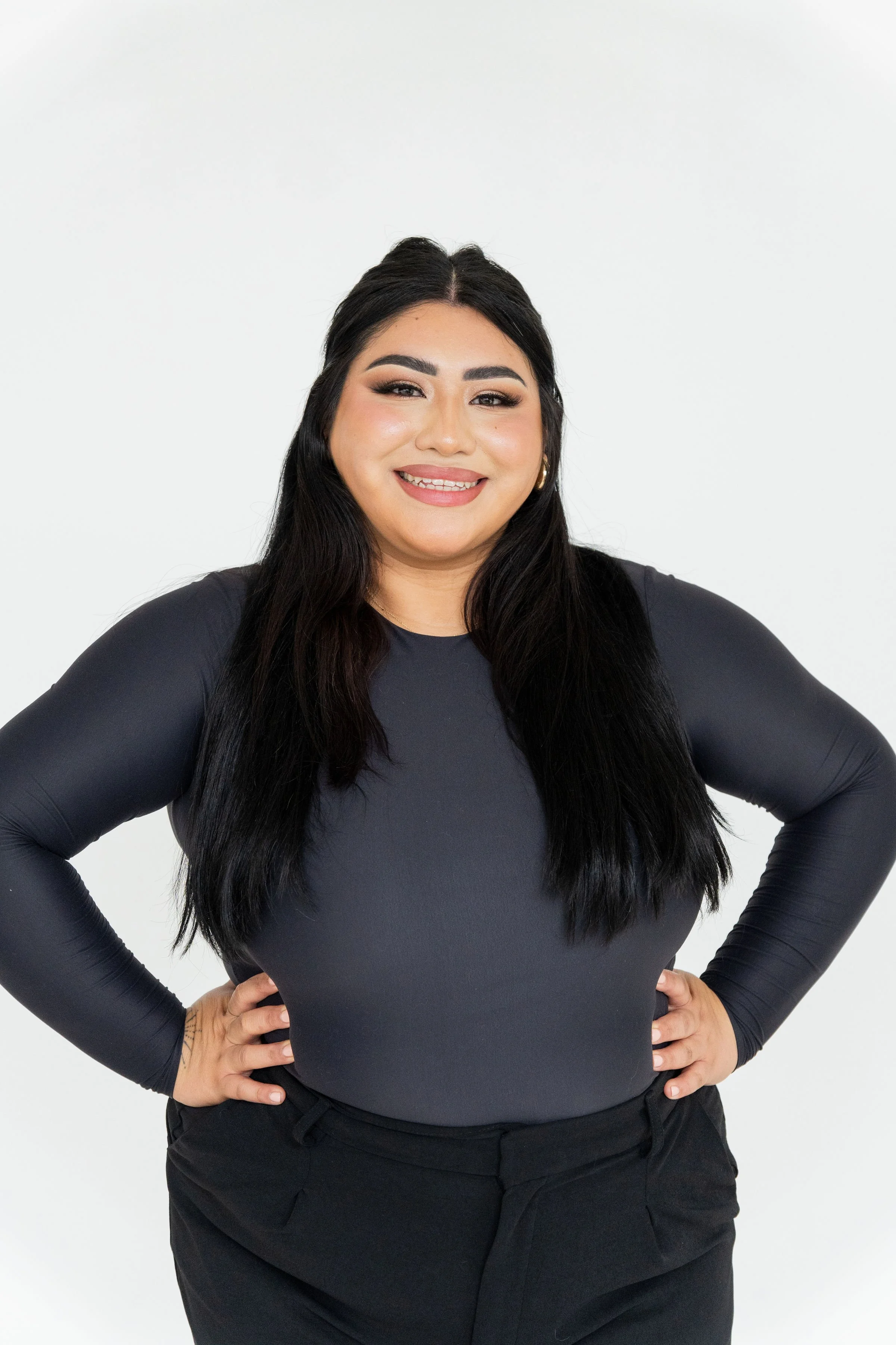 A woman with long black hair, wearing a black long-sleeve top and black pants, smiling with hands on hips against a plain white background.