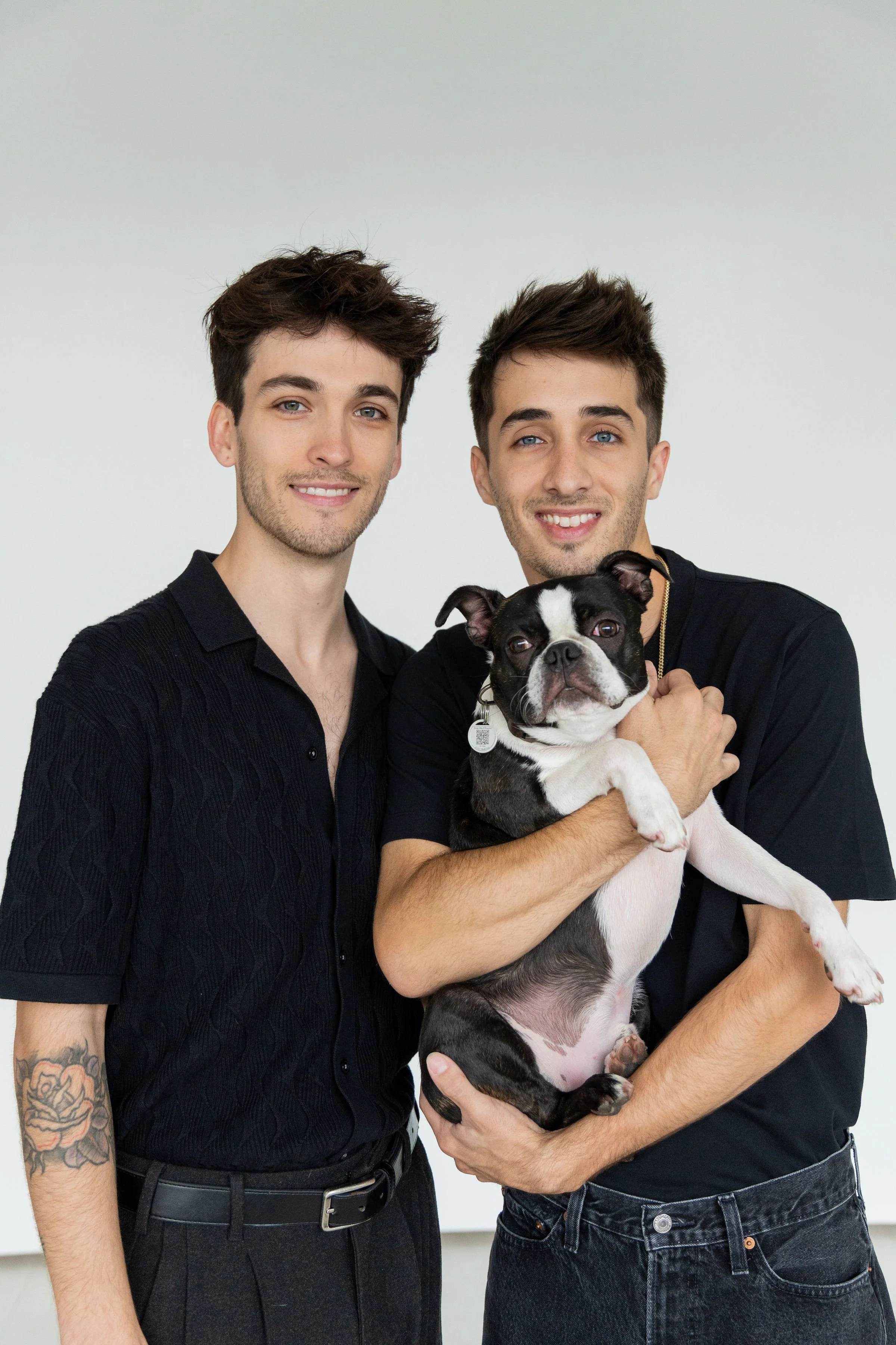 Two young men smiling, one holding a Boston Terrier dog, in front of a plain white background.