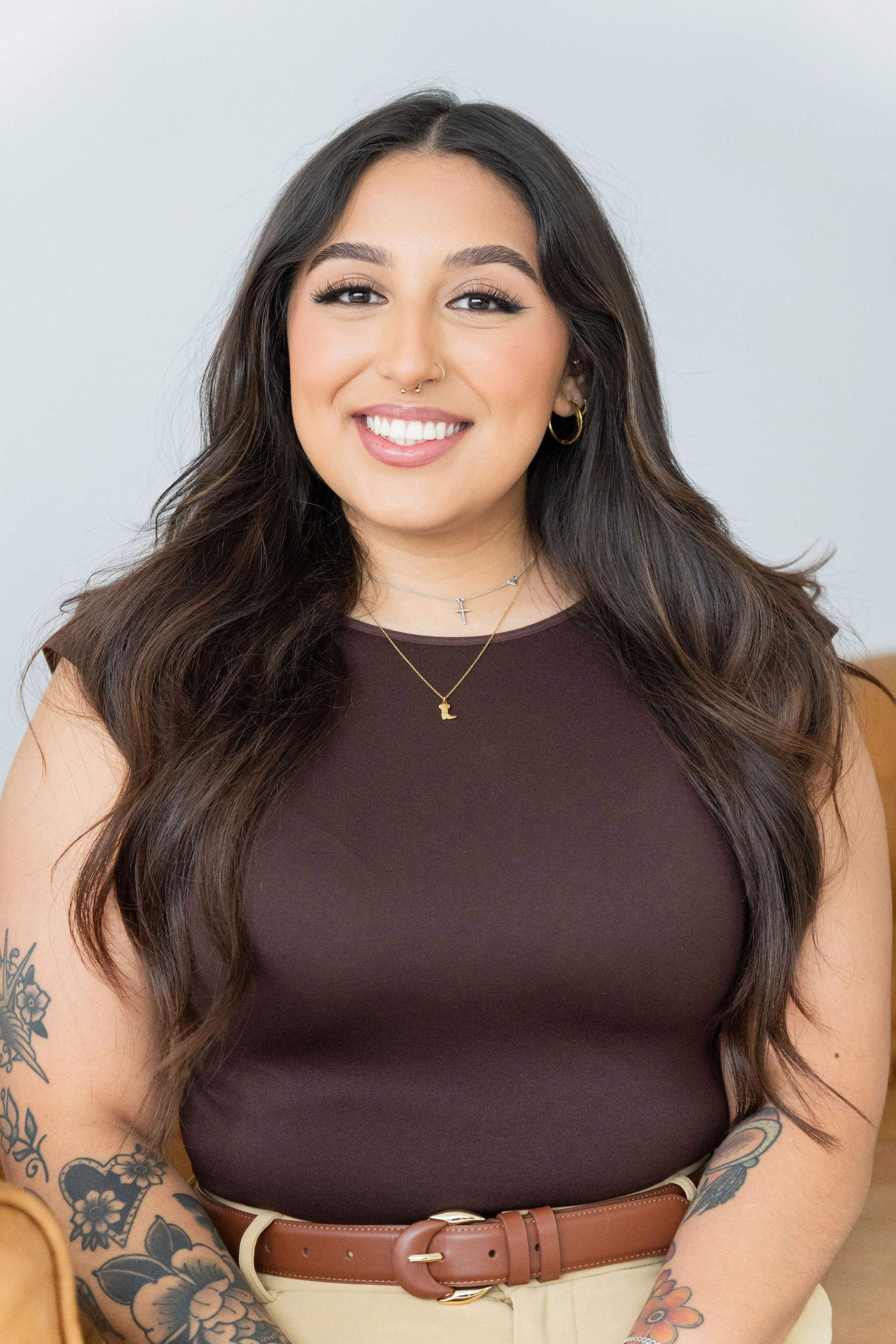A woman with long dark hair smiling at the camera, wearing a sleeveless dark top, layered necklaces, hoop earrings, and visible tattoos on her arms.