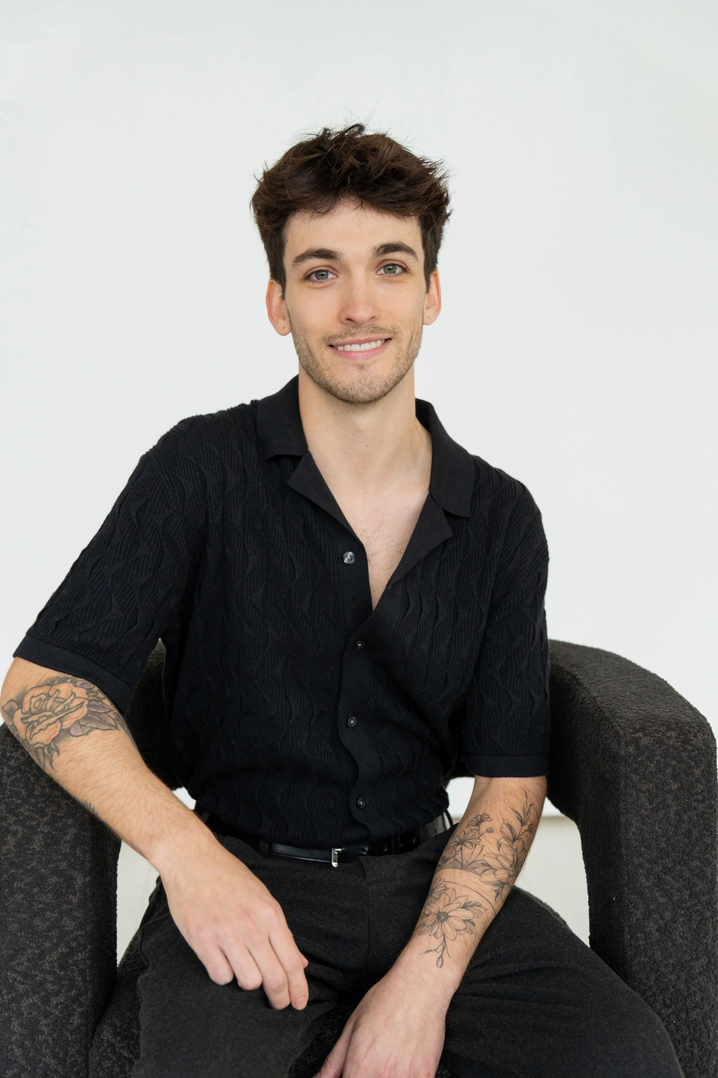 A young man with brown hair, blue eyes, and light skin, smiling while seated in a dark armchair against a plain white background, wearing a black short-sleeved button-up shirt and black pants, with tattoos on both arms.