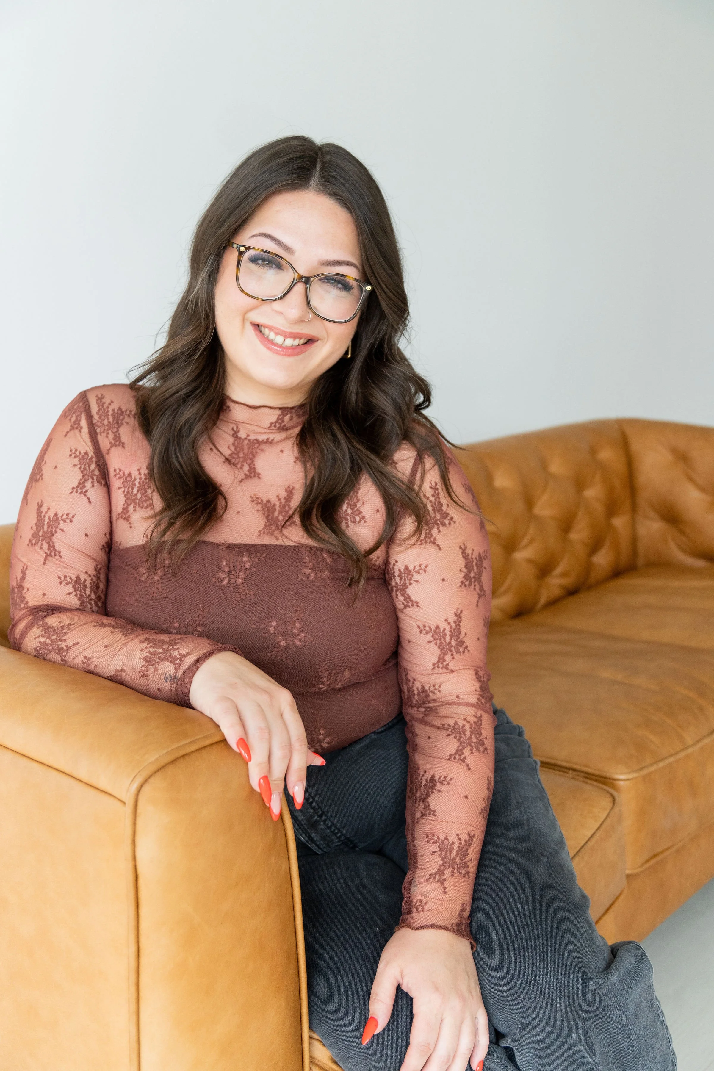 A woman with long dark hair, glasses, and red-painted nails sitting on a tan leather couch, smiling at the camera, wearing a sheer brown lace top and dark jeans.