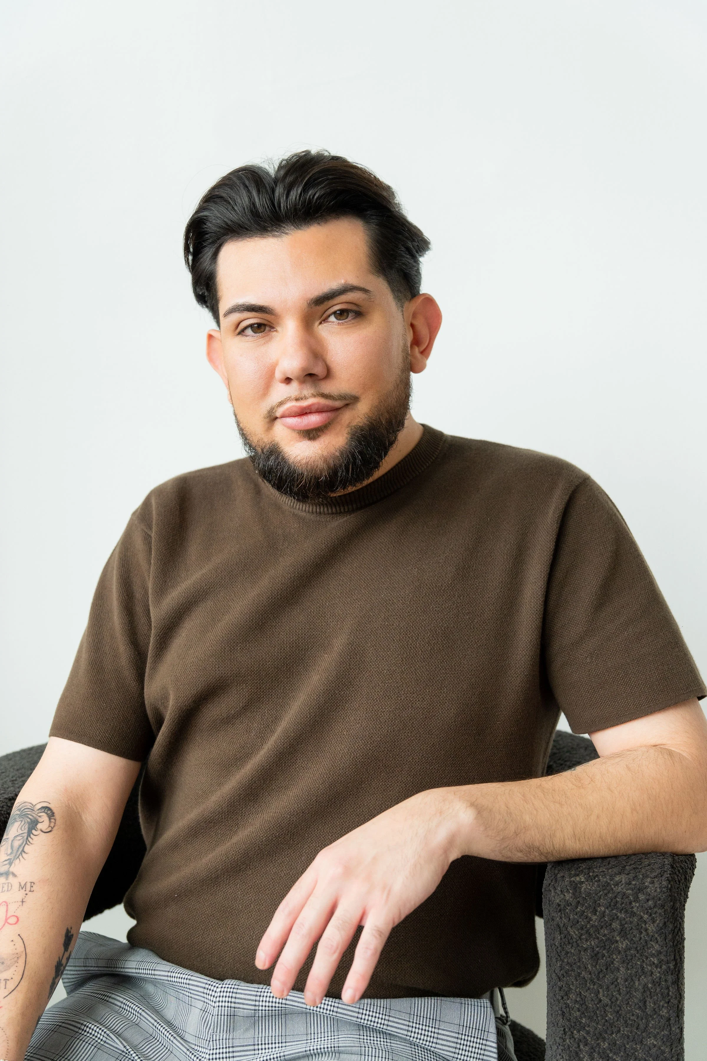 A young man with medium-length dark hair and a beard sitting on a dark gray chair against a plain white background, wearing a brown short-sleeved shirt and checkered pants.