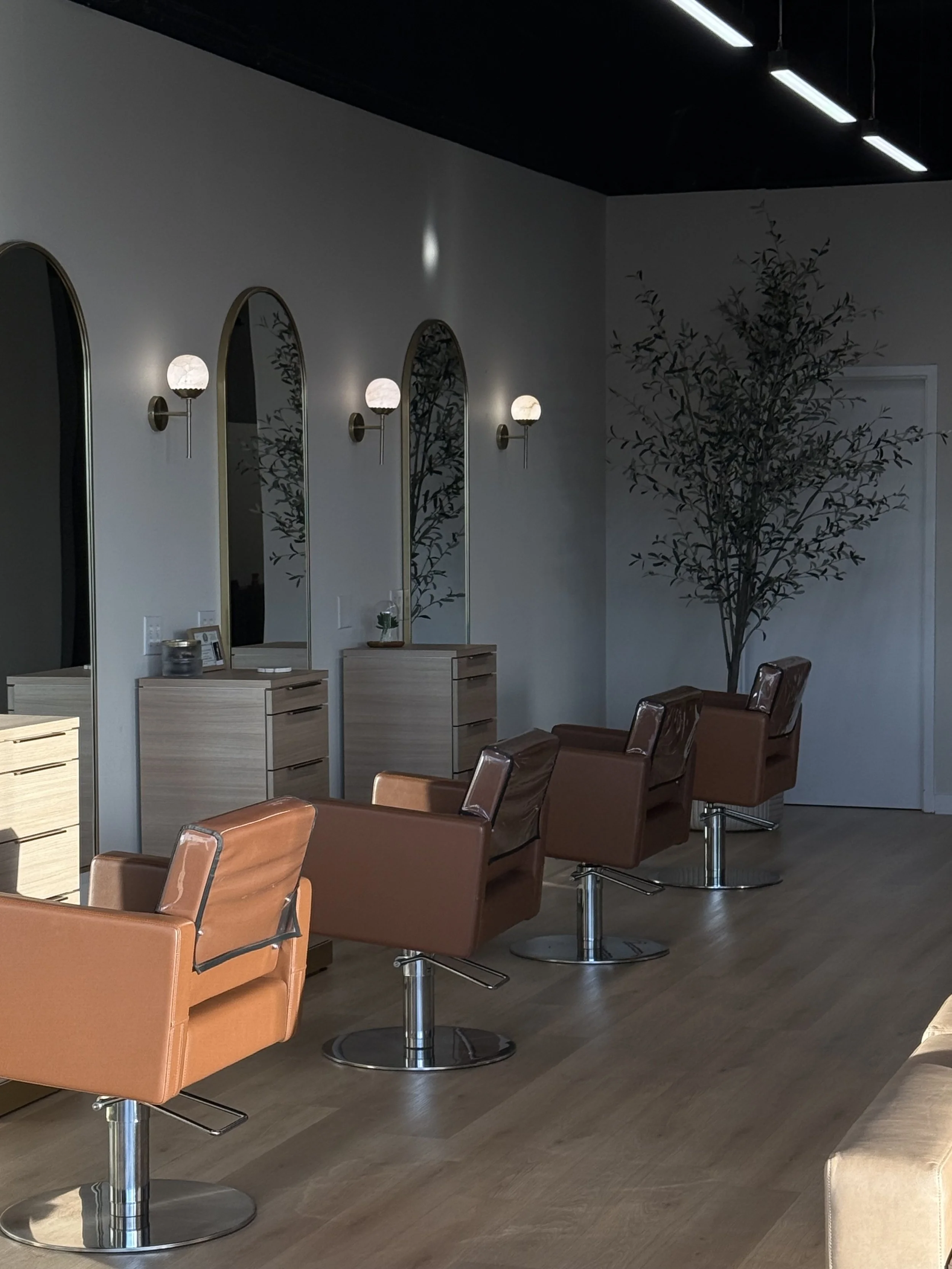 Empty salon with four brown styling chairs in front of three mirrors with wall sconces, a large tree in the corner, and wooden flooring.