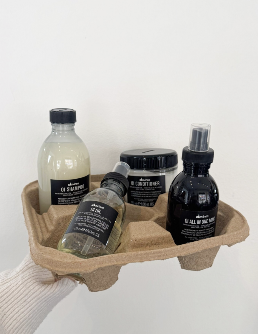 A biodegradable tray holds four black labeled containers of hair care products, including shampoo, oil, conditioner, and all-in-one milk.