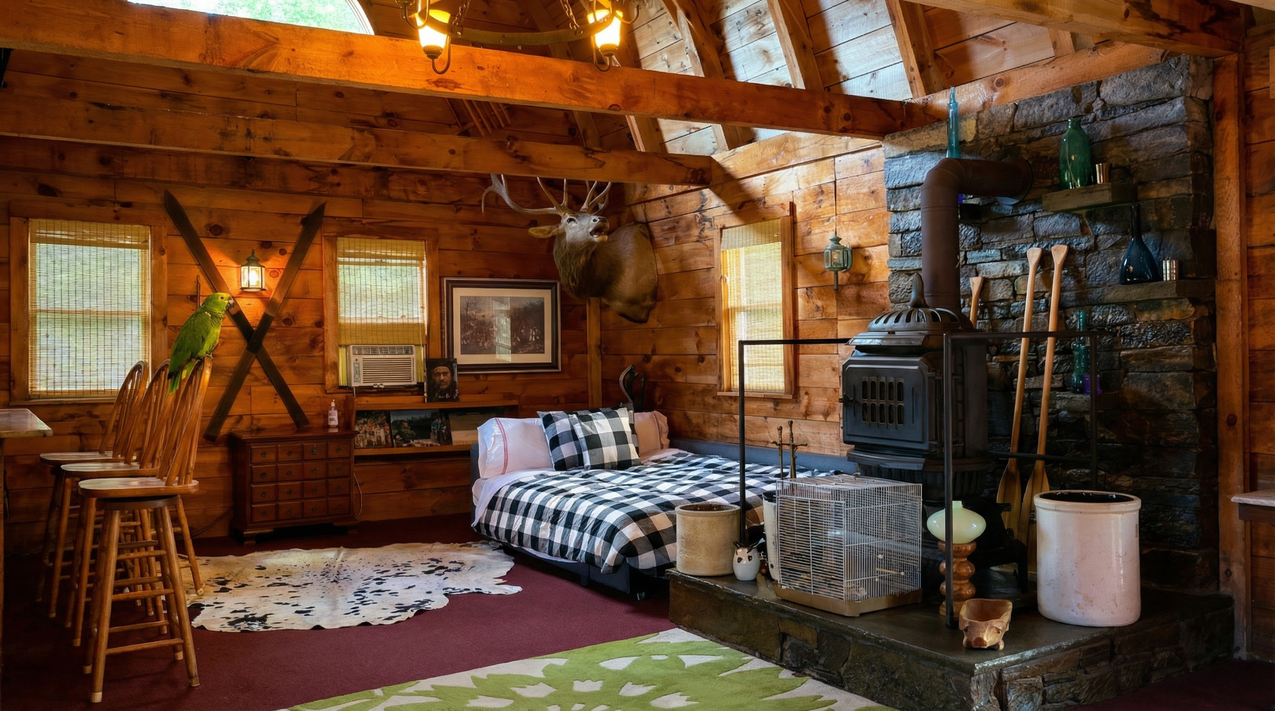 Cozy rustic cabin interior with a wood stove, mounted animal head, checkered bedding, parrot, and decorative items.