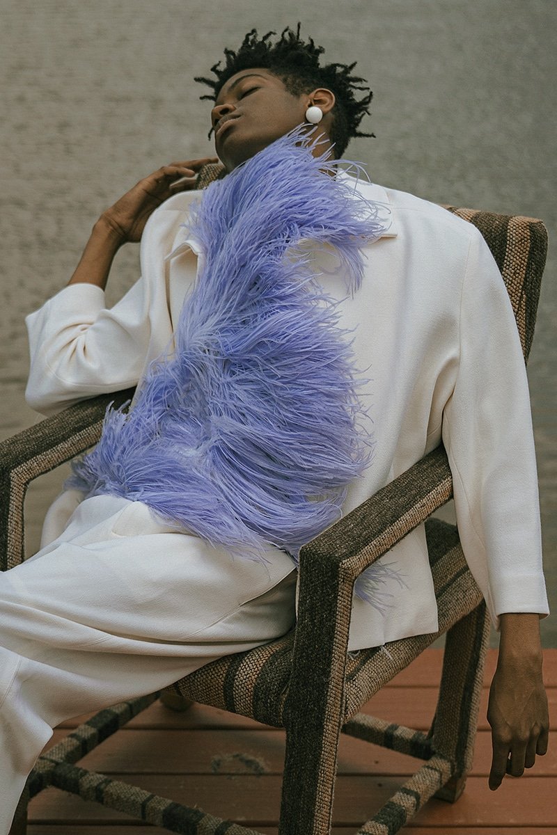 A woman relaxing in a woven chair, dressed in a white outfit with a purple feathered accessory draped over her shoulder. She has short, curly hair and is wearing white earrings, with her eyes closed and a peaceful expression.