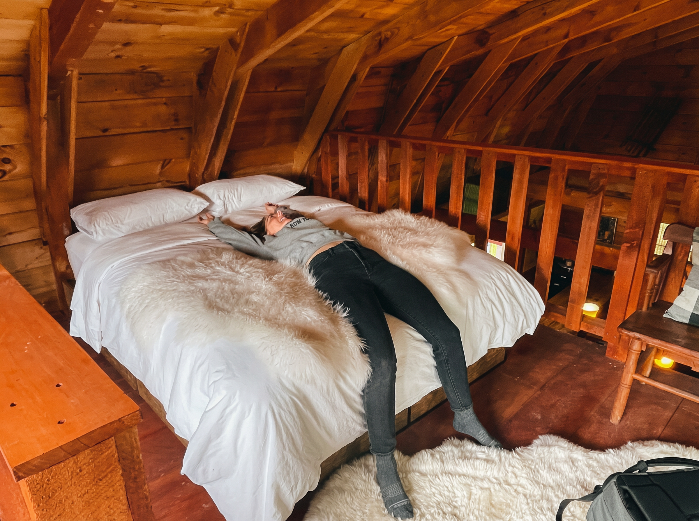 A woman lying on a bed with her arms stretched out in a cozy wooden room, wearing a sweatshirt and jeans.
