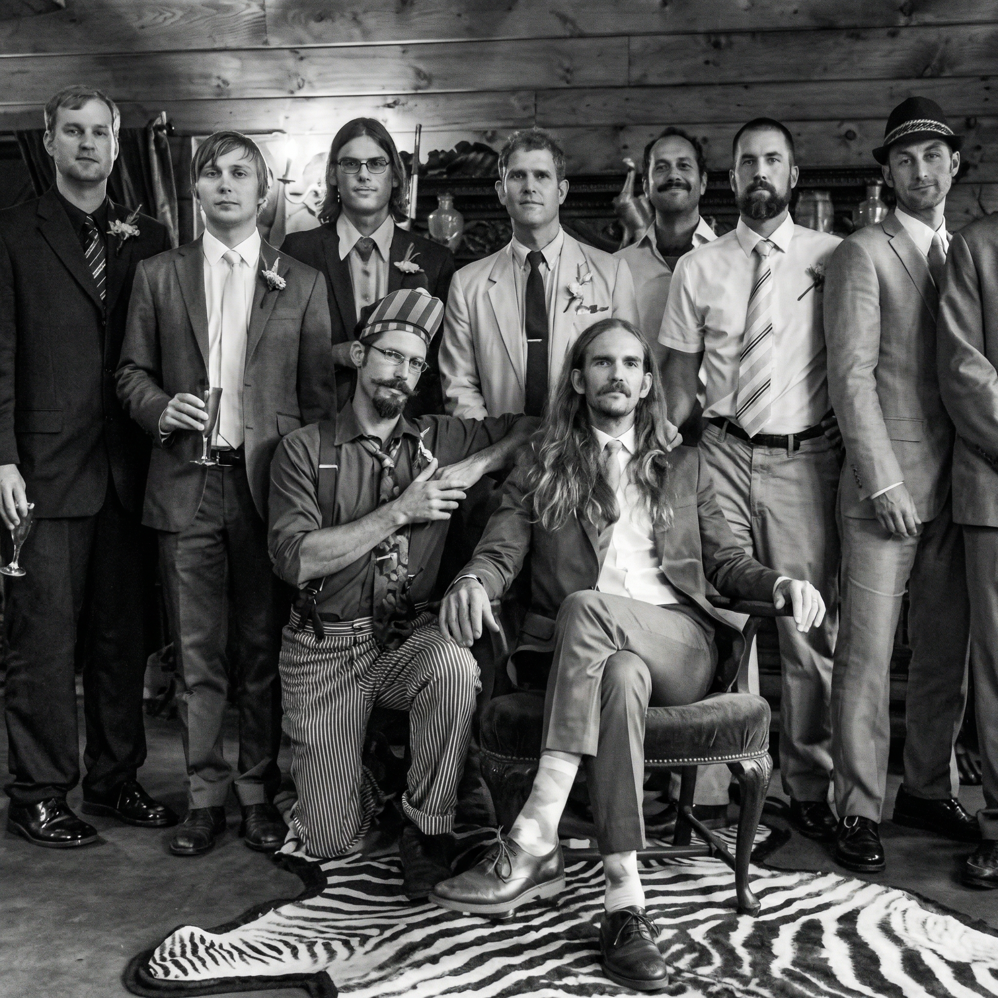 A group of men dressed in formal and vintage attire, some holding drinks, gathered in a rustic wooden interior.