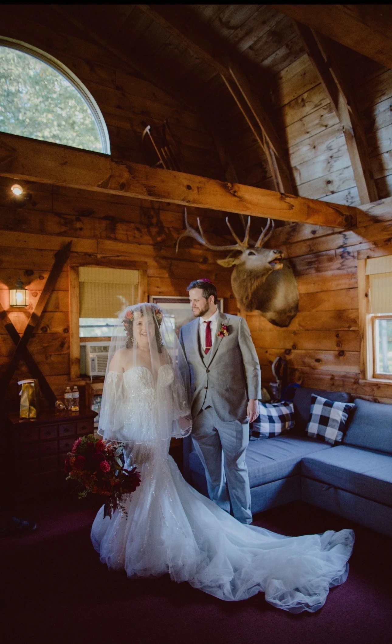 A bride and groom standing inside a rustic wooden cabin, with a large mounted deer head on the wall behind them. The bride is wearing a wedding dress with a veil and holding a bouquet, while the groom is in a gray suit with a red tie and boutonniere.