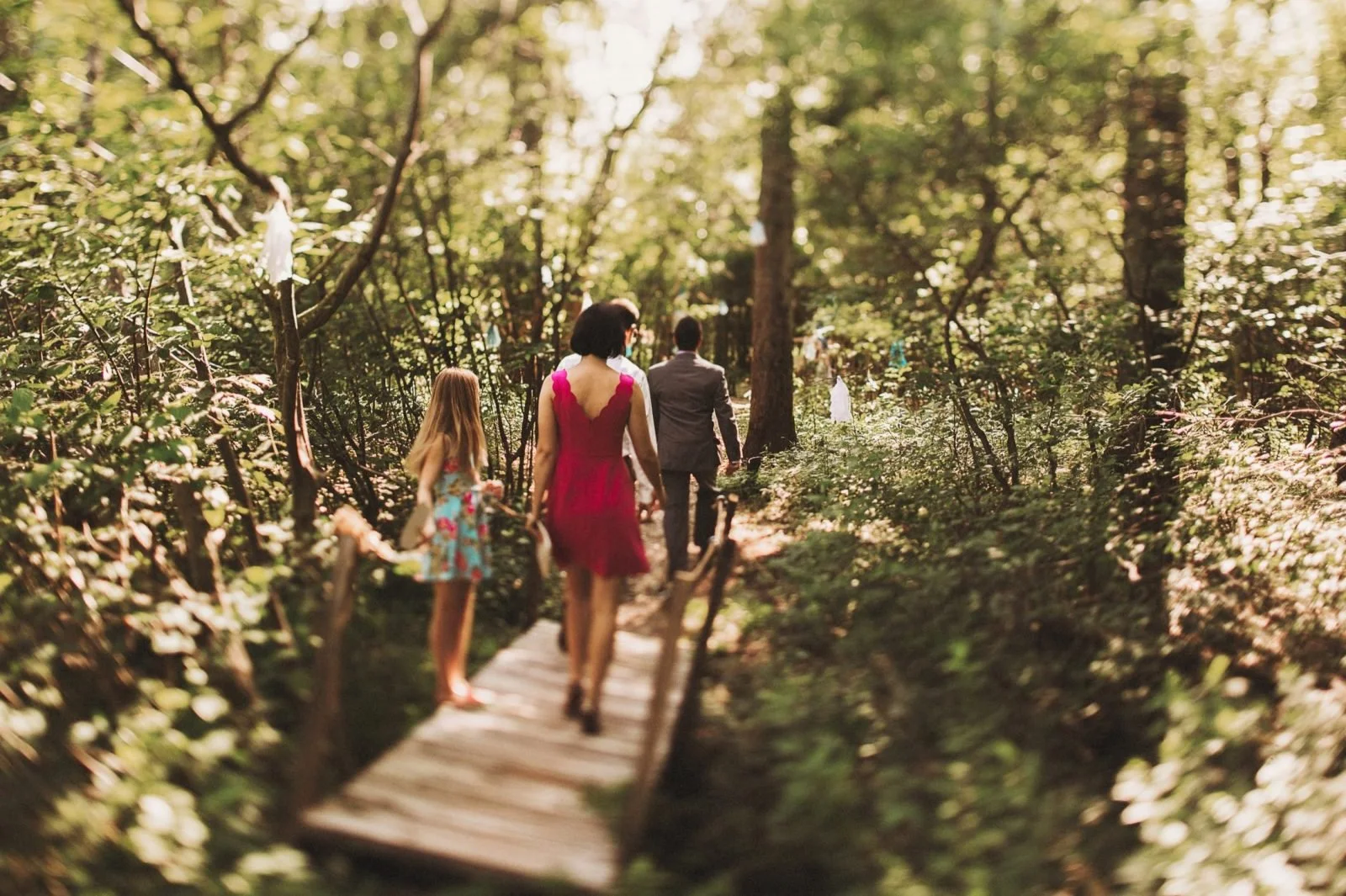 A woman, a man, and a girl are walking on a narrow wooden bridge through a lush green forest during daylight. The woman is wearing a red dress, the girl is wearing a colorful dress, and the man is in a suit.