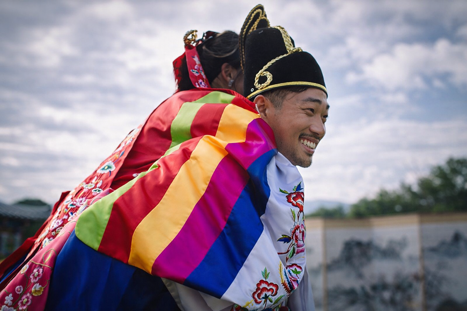 Two people in traditional Korean attire, one is being carried on the other's back, wearing a black hat with gold embroidery and a colorful rainbow silk sash. They are smiling with an outdoor background under a cloudy sky.