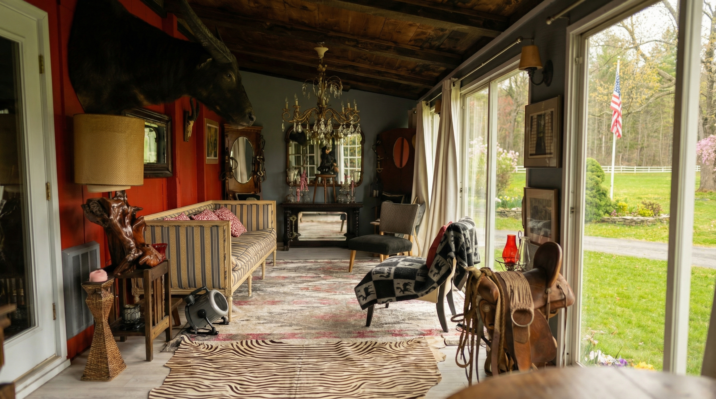 A cozy living room with eclectic decorations including animal-themed accents like a mounted boar head and a zebra print rug, large windows with cream curtains revealing a lush green yard, antique furniture, a chandelier, and various decorative items.