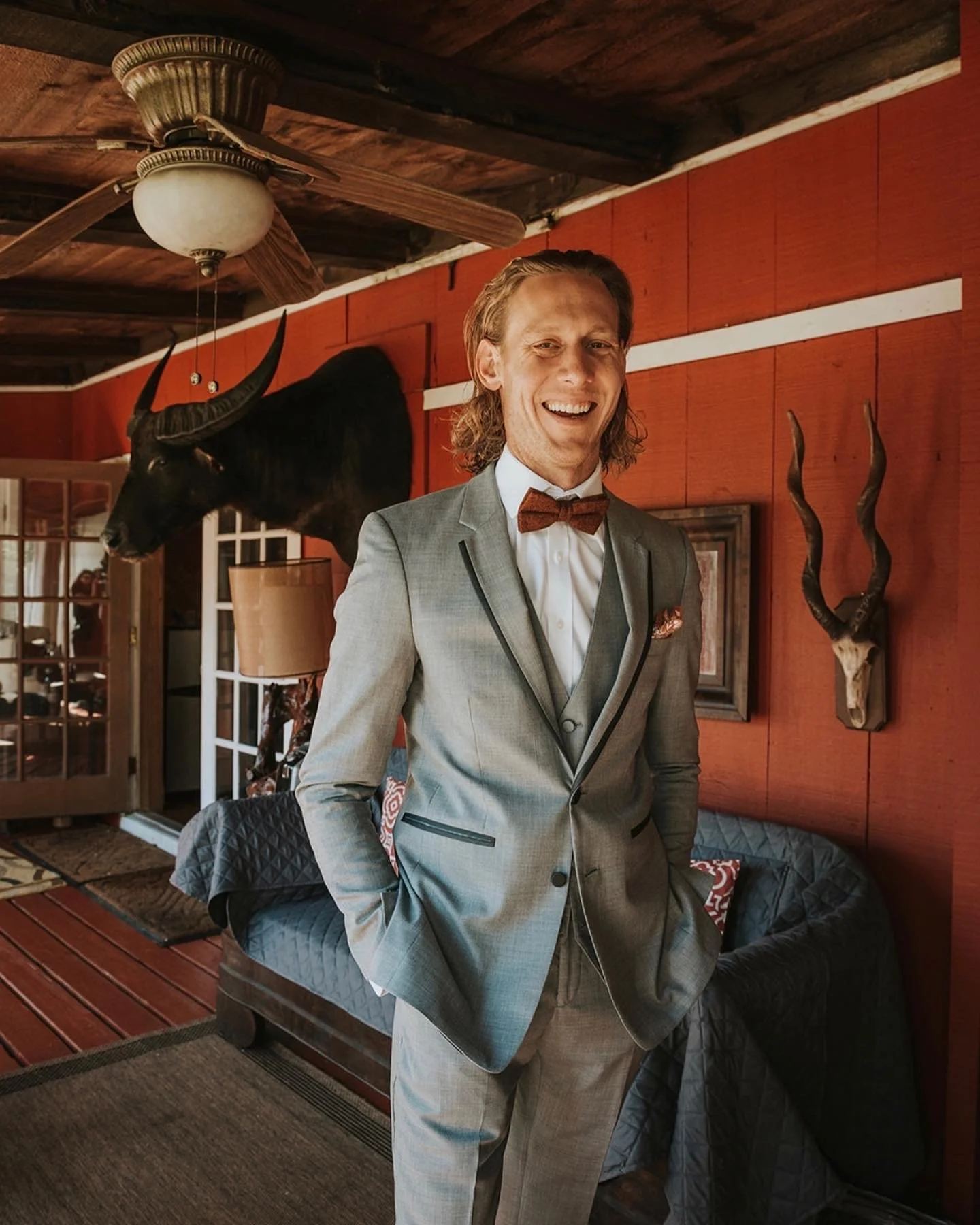 A young man in a gray suit with a bow tie, smiling and standing in a cozy, rustic room with hunting-themed decorations, including mounted animals on the red wall.