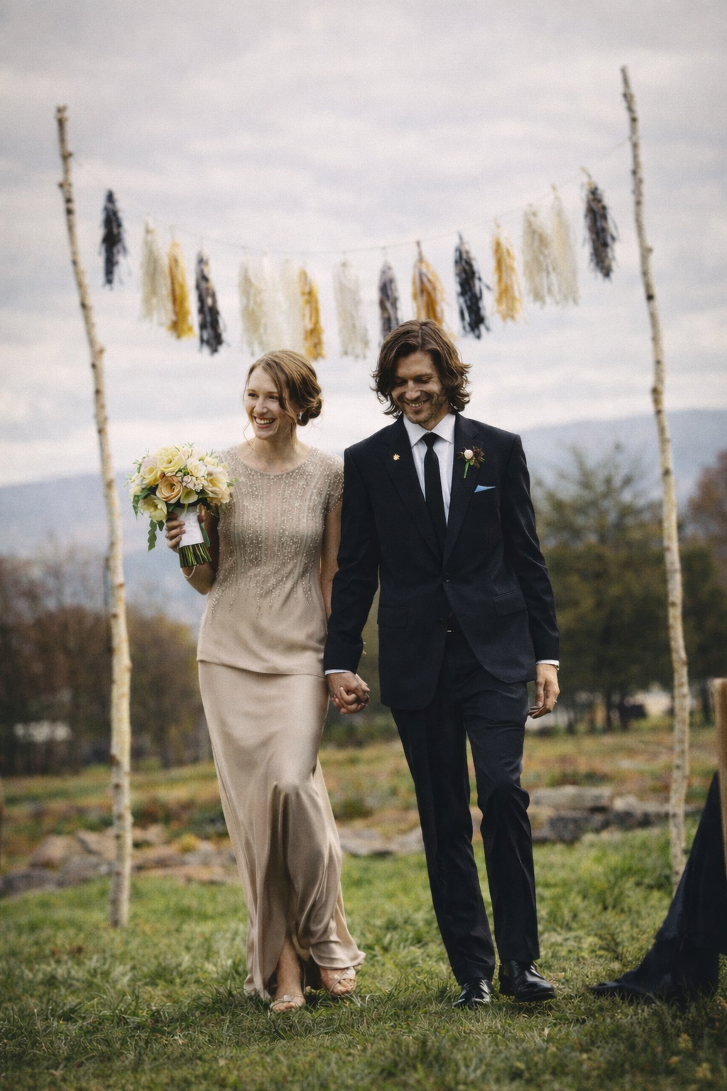 A newlywed couple walking hand in hand outdoors under a decorated arch, with a woman in a beige dress holding a bouquet, and a man in a dark suit, smiling.