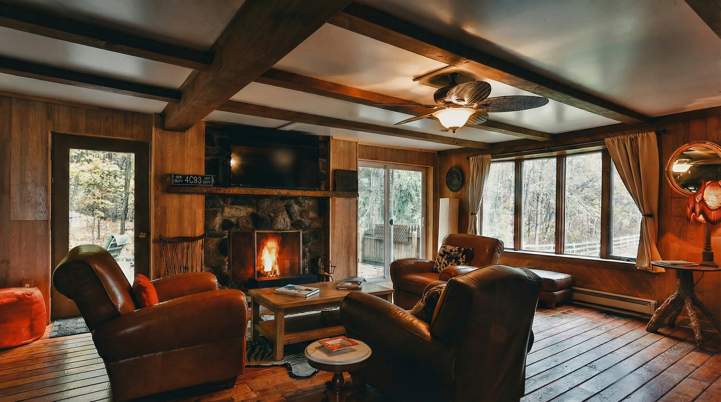 Living room with wood-paneled walls, large windows, leather armchairs, a fireplace with a fire, a wooden coffee table, and a flat-screen TV mounted above the fireplace.