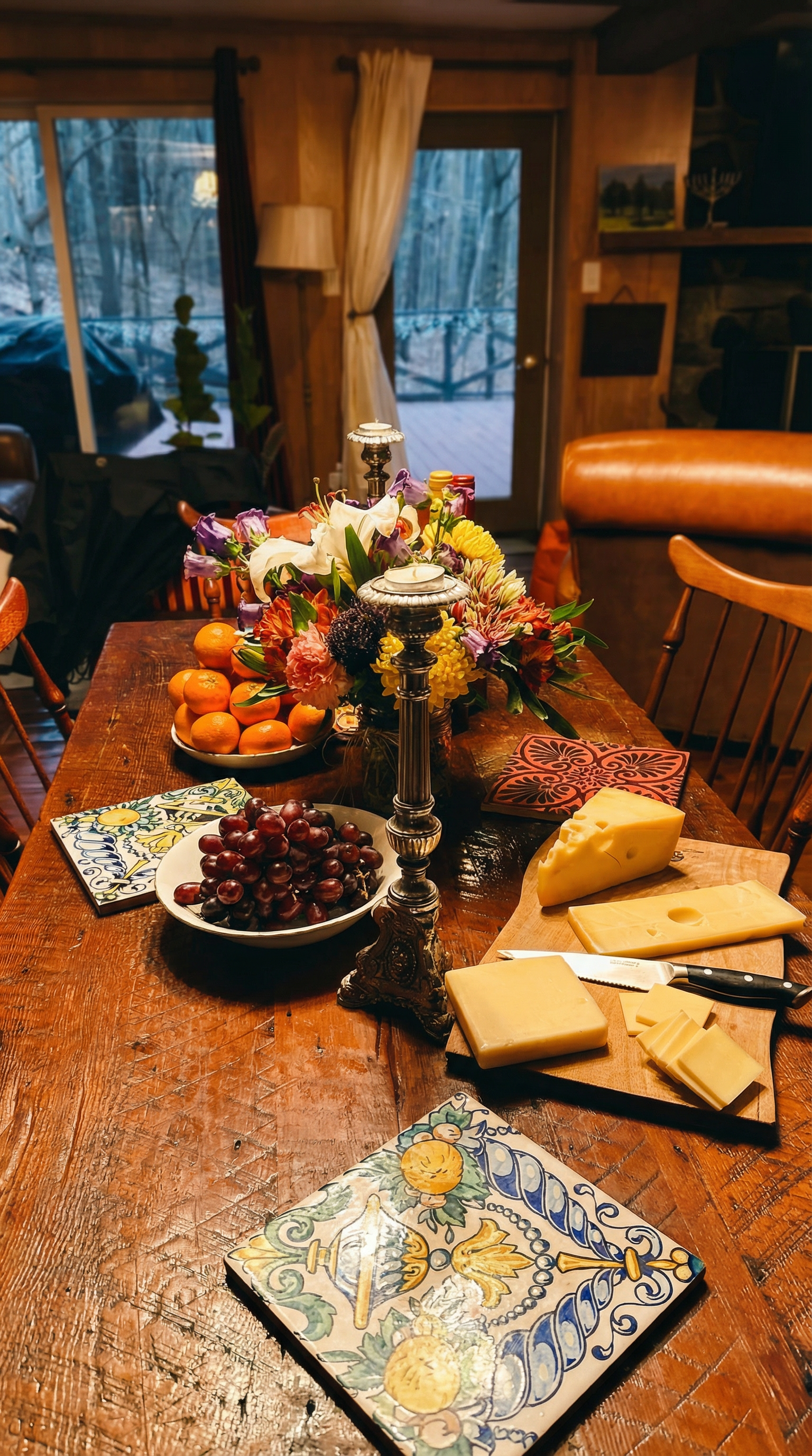 A rustic wooden dining table set with a colorful flower arrangement, a plate of red grapes, a bowl of oranges, and imported cheeses and a knife. The background shows a cozy room with wood-paneled walls, a sliding glass door leading outside, and warm indoor lighting.