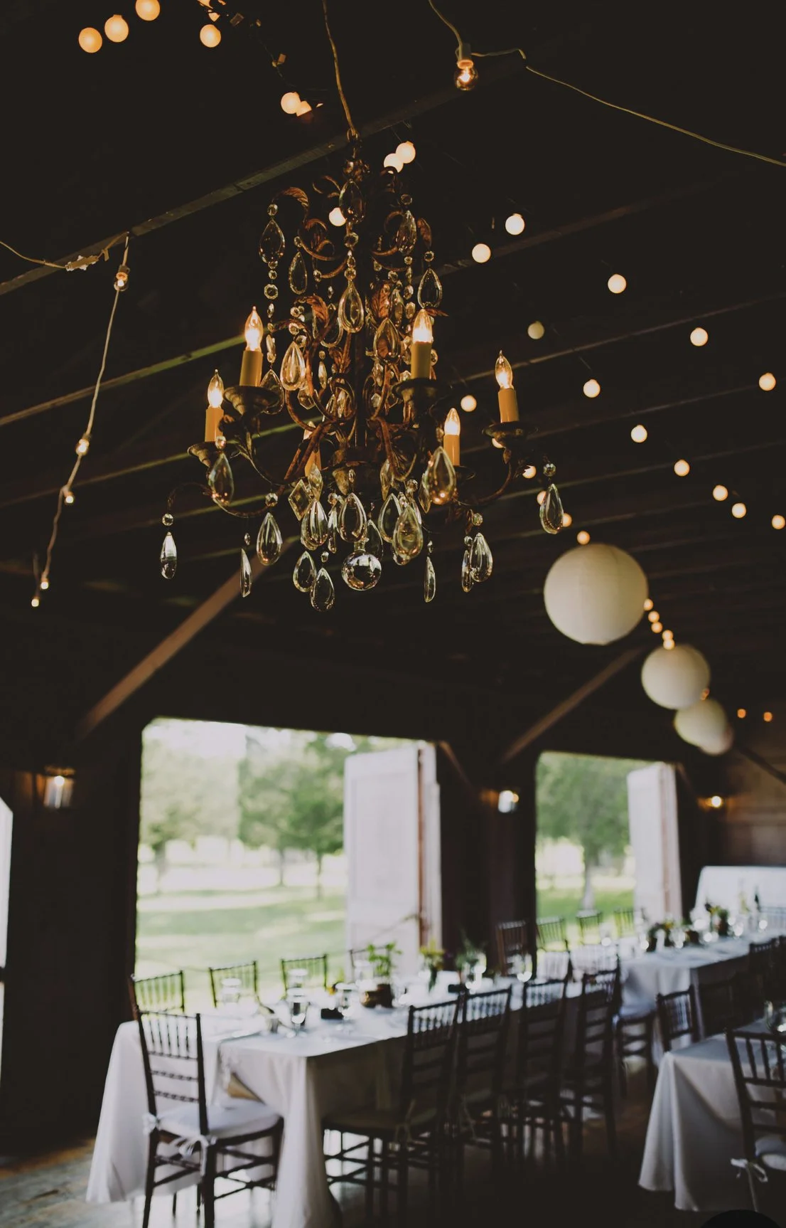 Chandelier hanging from the ceiling with string lights and paper lanterns, over a decorated dining table in a rustic indoor setting.
