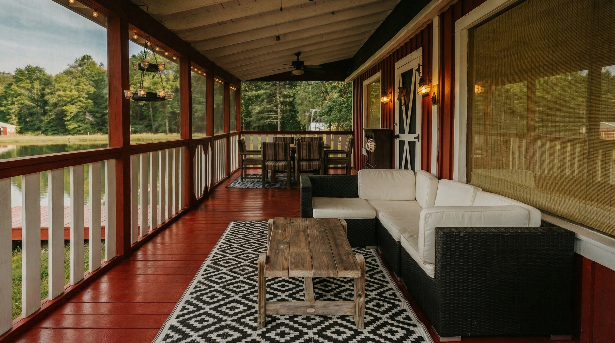 Covered porch with white ruffled curtains, red wooden flooring, black wicker sofa with white cushions, rustic wooden coffee table, black and white geometric rug, dining table with six striped chairs, lake view through windows, and warm outdoor lighting.