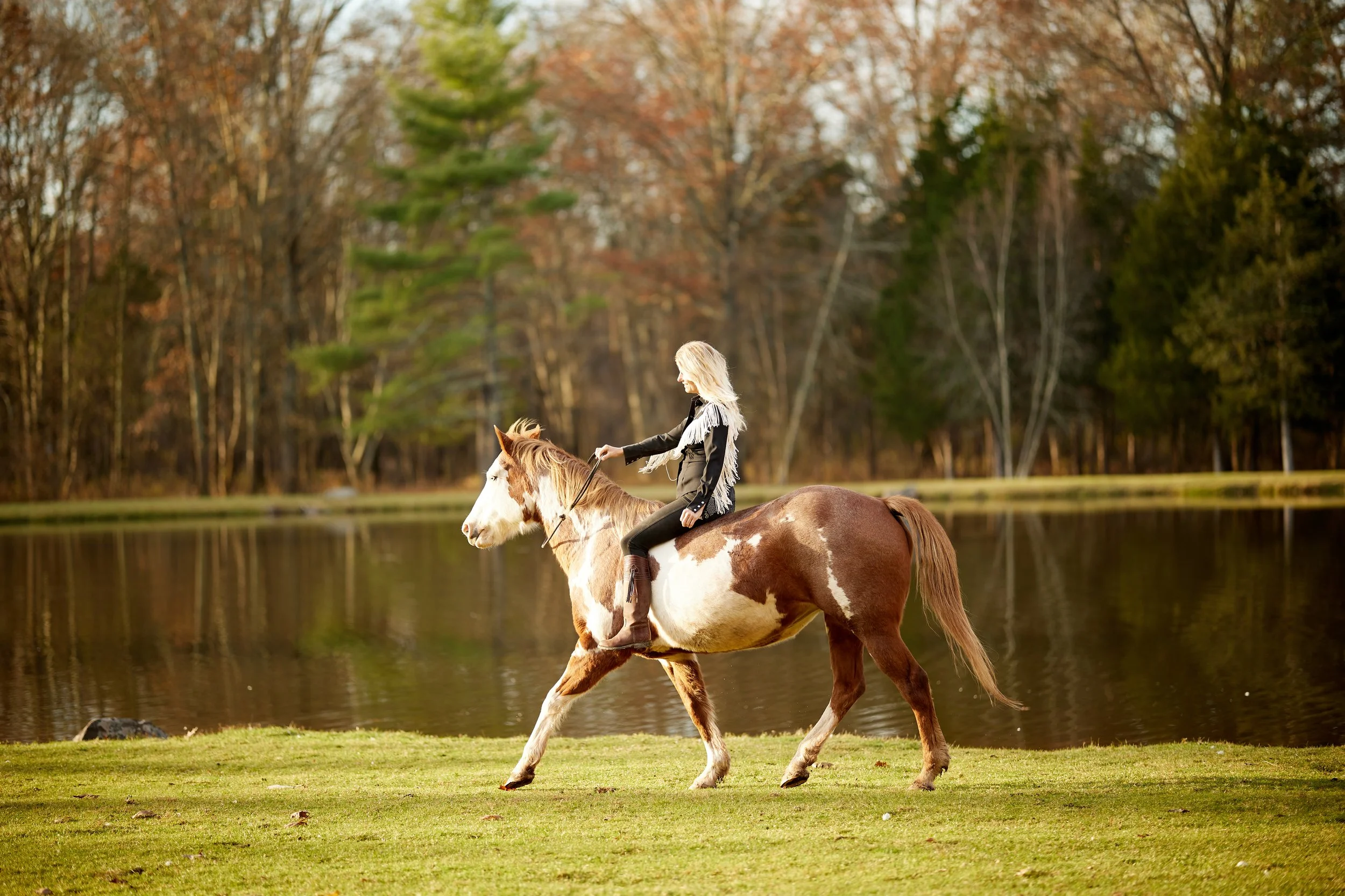 Woman riding a brown and white pinto horse near a pond with autumn-colored trees in the background.