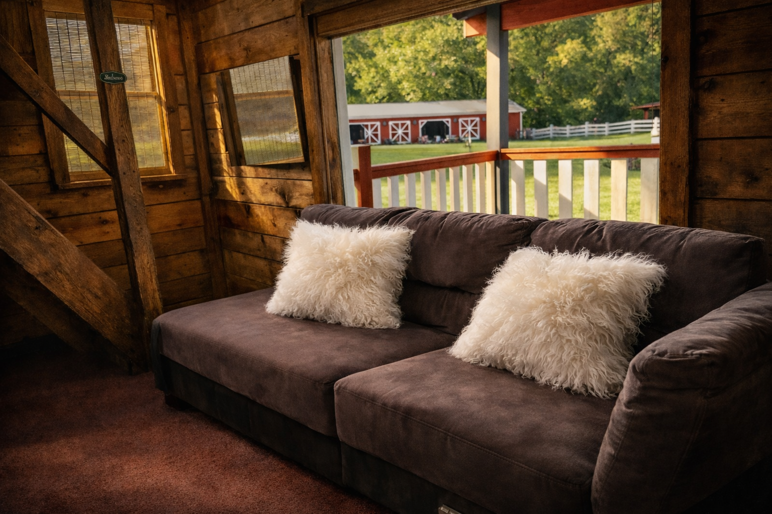 A rustic wooden interior with a brown sofa decorated with two fluffy white pillows, overlooking a farm scene with a red barn, white fence, and green trees outside.