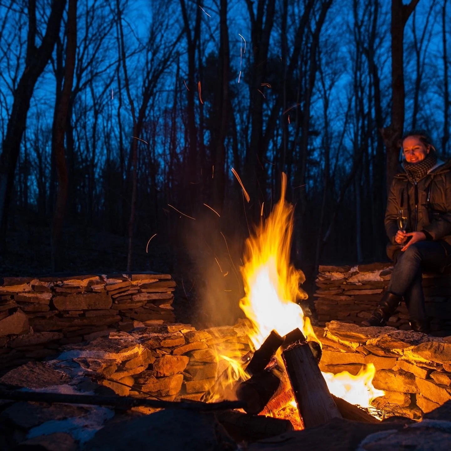 Person sitting by a campfire in a wooded area at dusk, smiling and holding a wine glass.