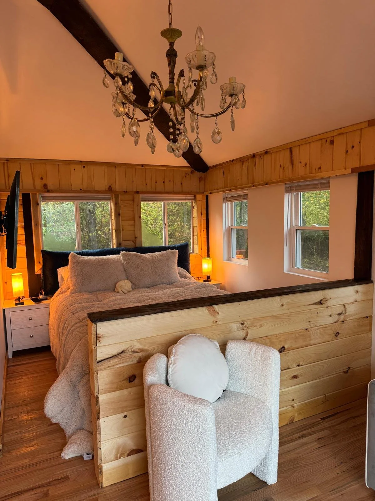A cozy bedroom with wooden walls and ceiling, a chandelier hanging from the ceiling, a bed with plush pillows and a blanket, side tables with lamps, and a white armchair in front of a wooden partition.