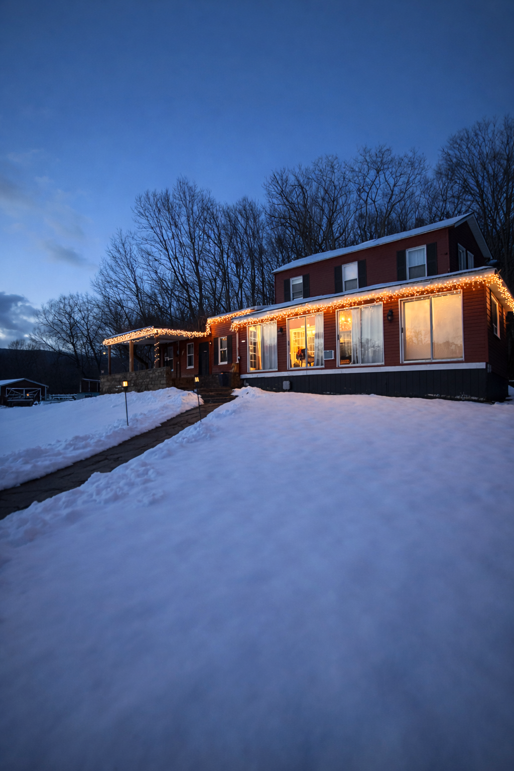 A house decorated with Christmas lights, surrounded by snow, during twilight with a dark blue sky and leafless trees in the background.
