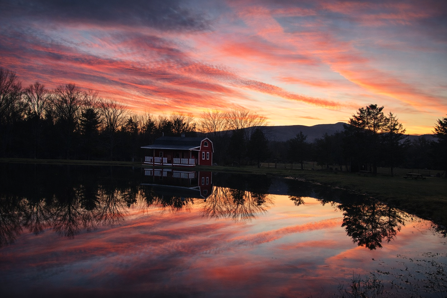 A peaceful landscape with a vibrant sunset sky and scattered clouds reflected on a still pond. Silhouettes of leafless trees, a red barn, and distant mountains are visible as the scene reflects on the water.