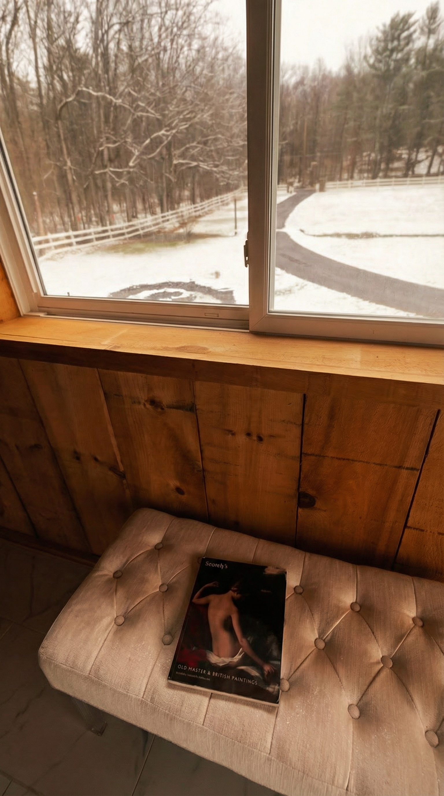 A beige tufted bench with a magazine on top, situated in front of a wooden wall and beneath a large window showing a snowy outdoor scene with trees and a winding pathway.