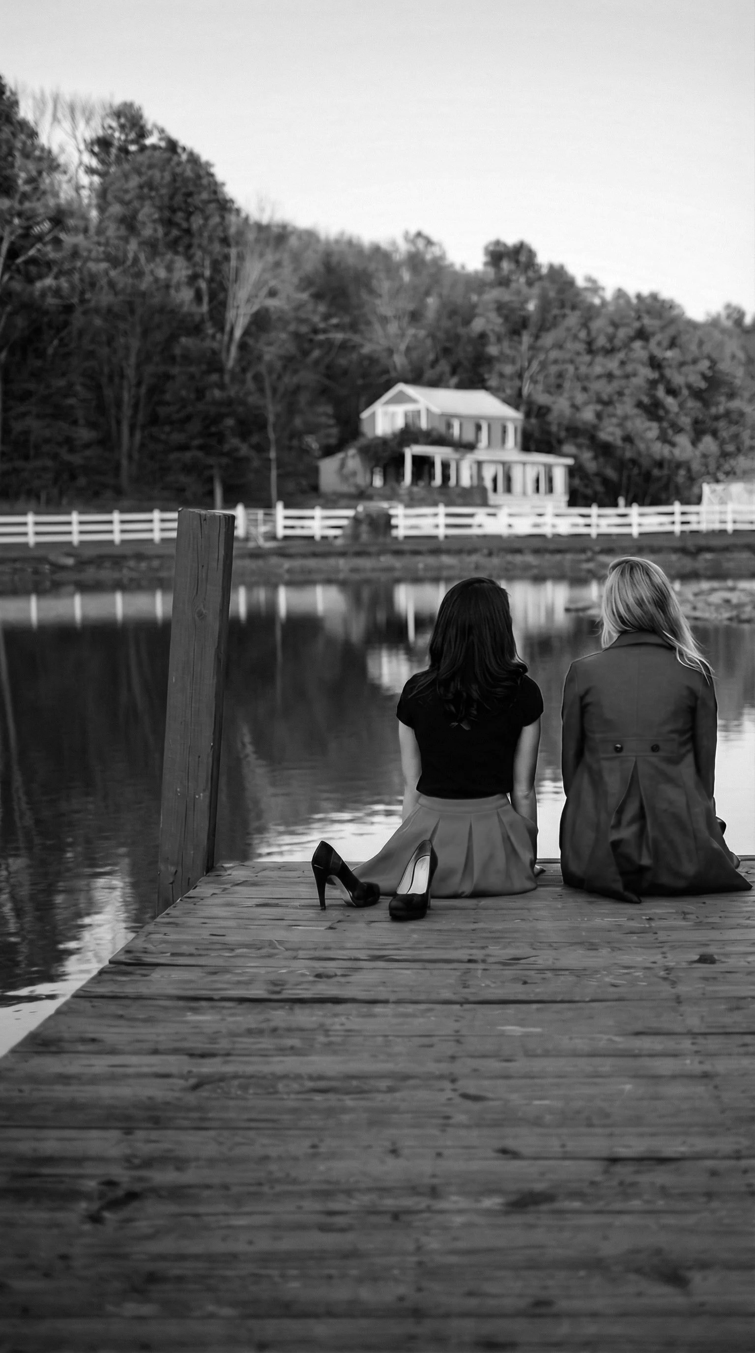 Two women sitting on a wooden dock by a lake, with high heels placed beside them, overlooking a house across the water.