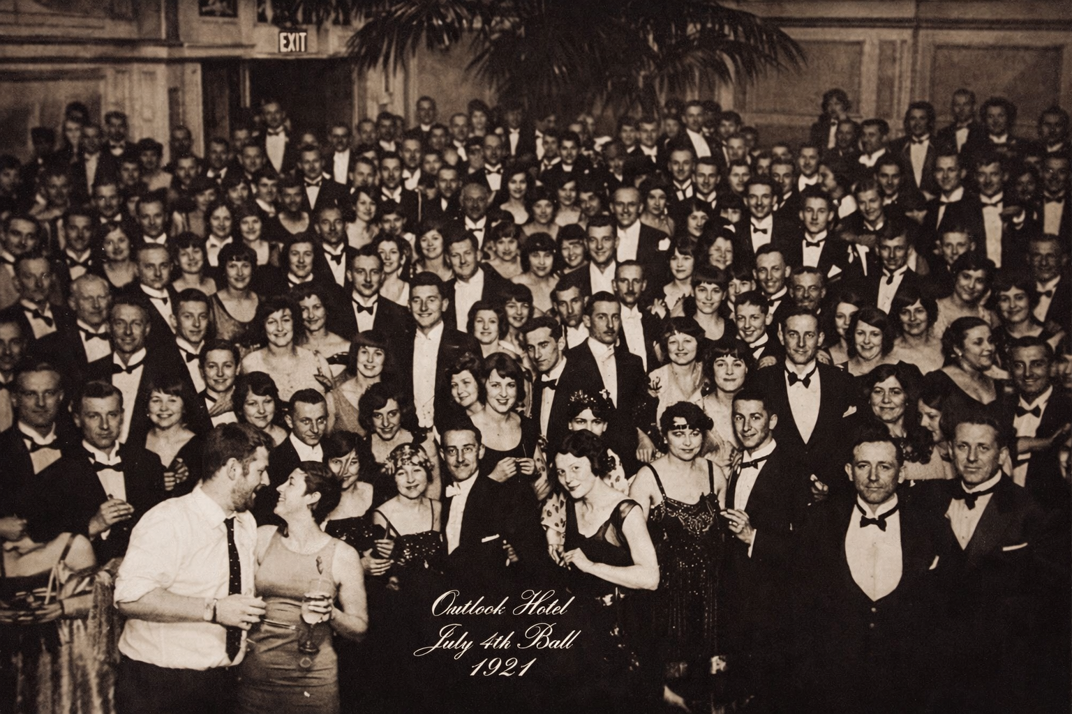 A large group of people dressed in formal attire for a 1921 July 4th ball at the Outlook Hotel, with many women in flapper dresses and men in tuxedos, gathered in a grand hall with a palm tree and an exit sign visible in the background.