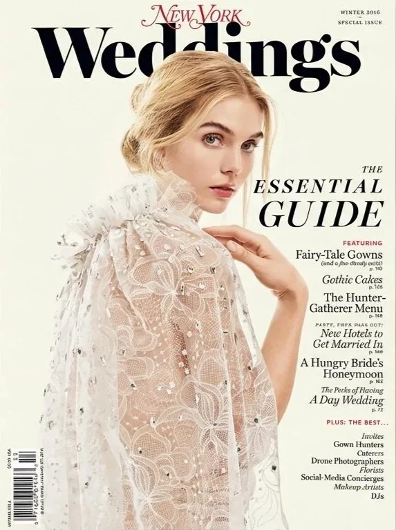 A woman with purple hair is dressed in a long brown designer coat with an orange dress underneath. She stands in a fenced pasture, facing away, under a cloudy sky.