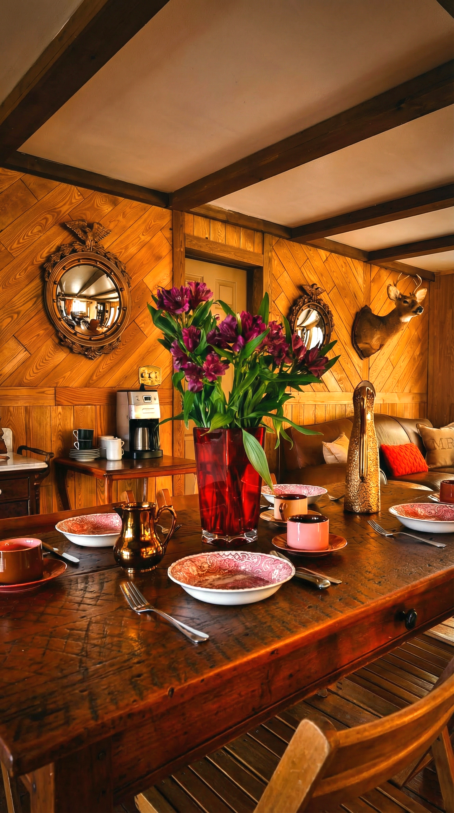 A cozy wooden dining room with a table set for a meal, featuring pink and white bowls, cups, and silverware. A large red vase with purple flowers is in the center. The wall has wood paneling with decorative mirrors and a mounted deer head, with a leather sofa and pillows in the background.